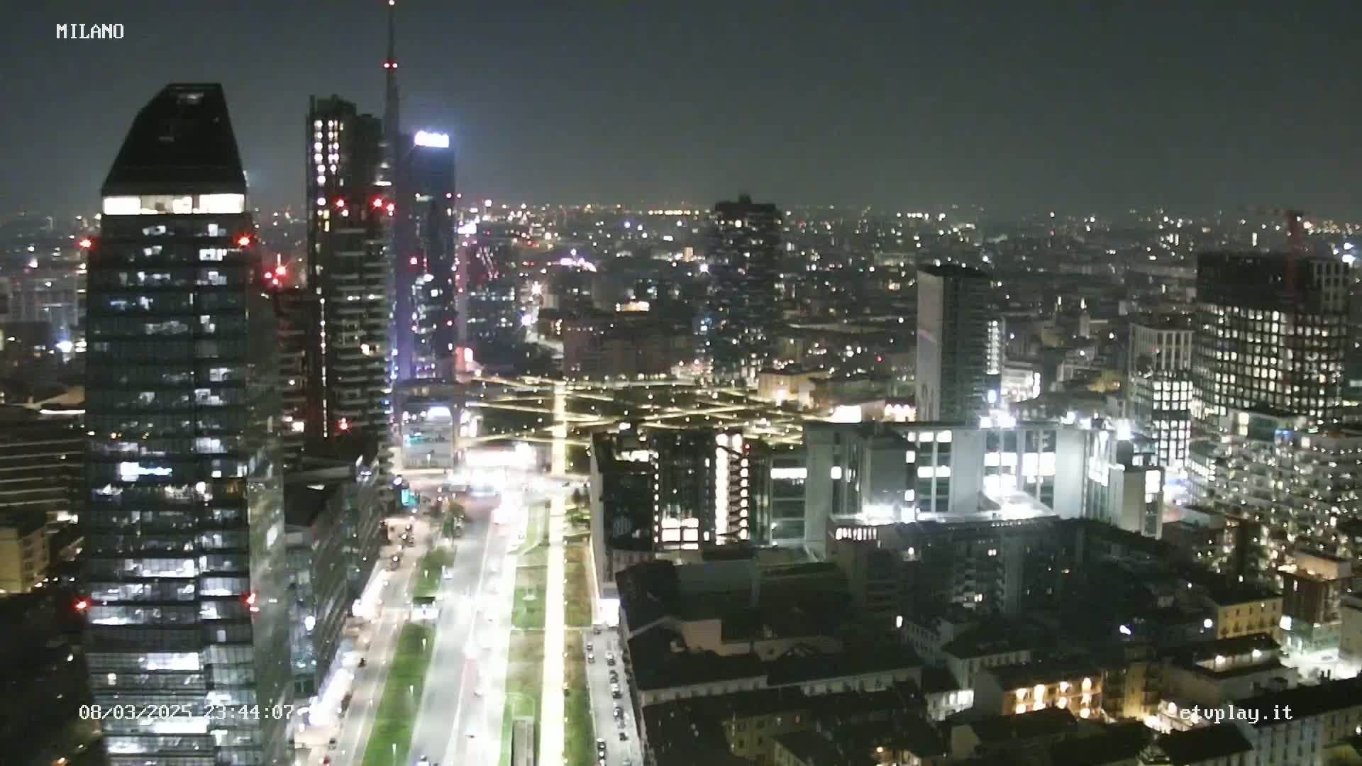 A nighttime aerial view of a city with numerous skyscrapers and brightly lit streets, under a clear, dark sky.