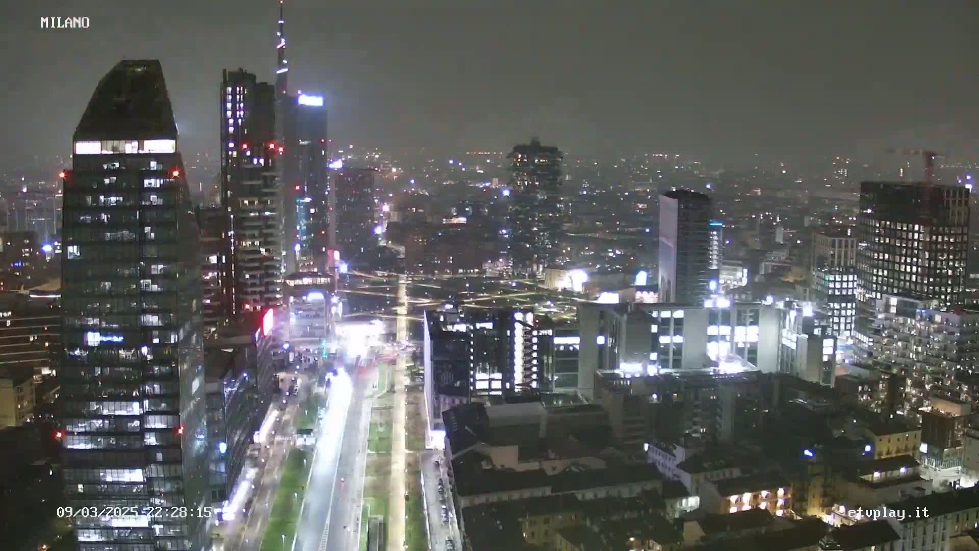 A nighttime, hazy view of a city skyline featuring numerous skyscrapers and brightly lit streets.