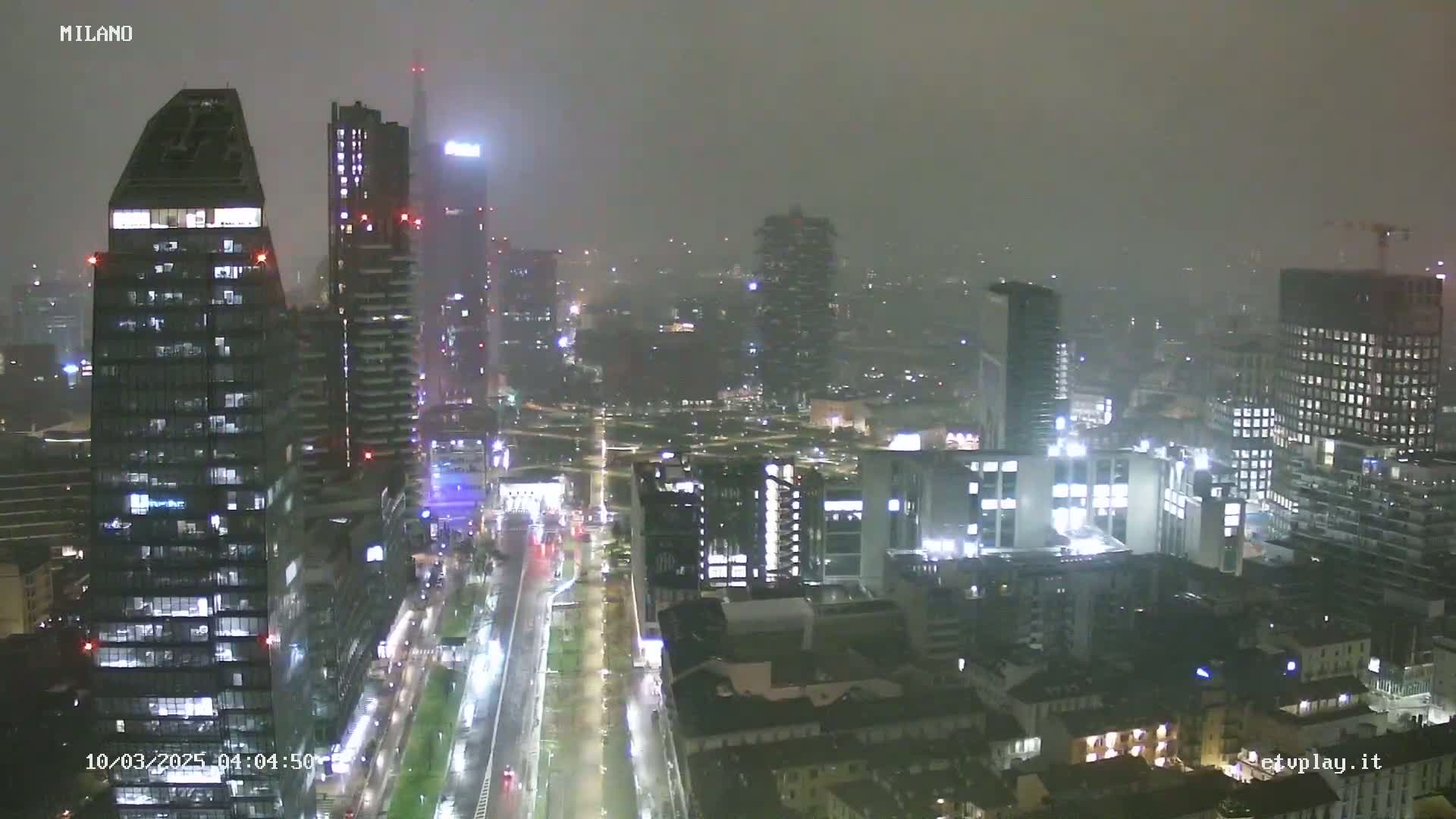 A nighttime view of a city skyline shrouded in heavy fog or mist, with illuminated buildings and streets below.