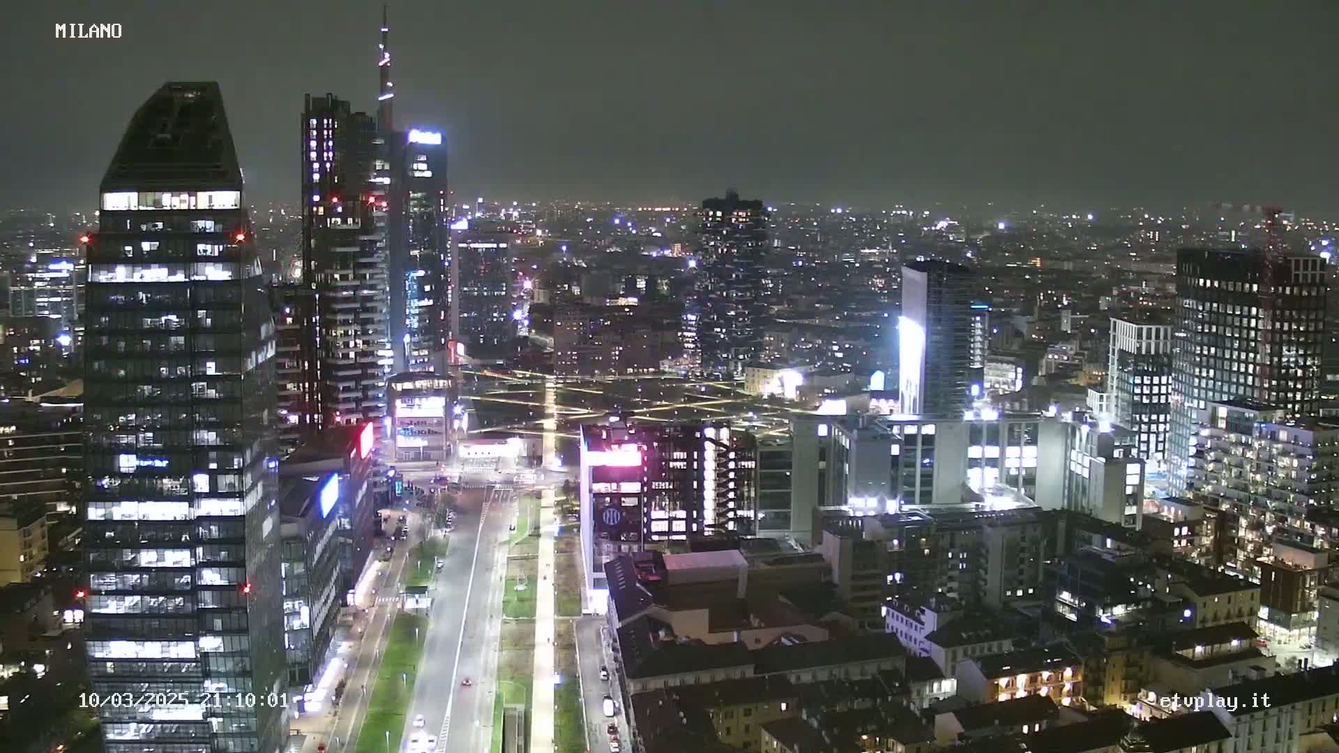 A nighttime, high-angle view of a city skyline with numerous illuminated skyscrapers and buildings, featuring a wide road below.