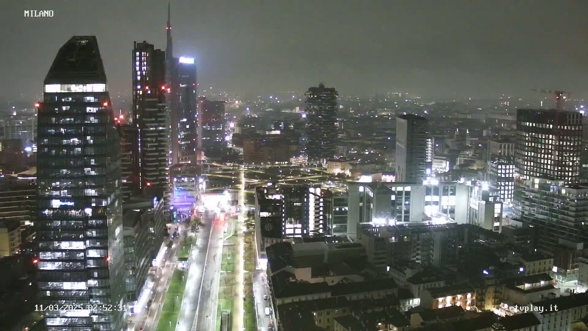 A nighttime view of a city skyline with many tall buildings illuminated, under a hazy, possibly rainy sky.