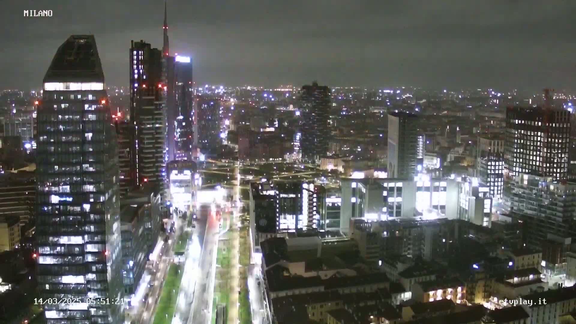 A nighttime aerial view of a city skyline with many illuminated buildings and a brightly lit street below.