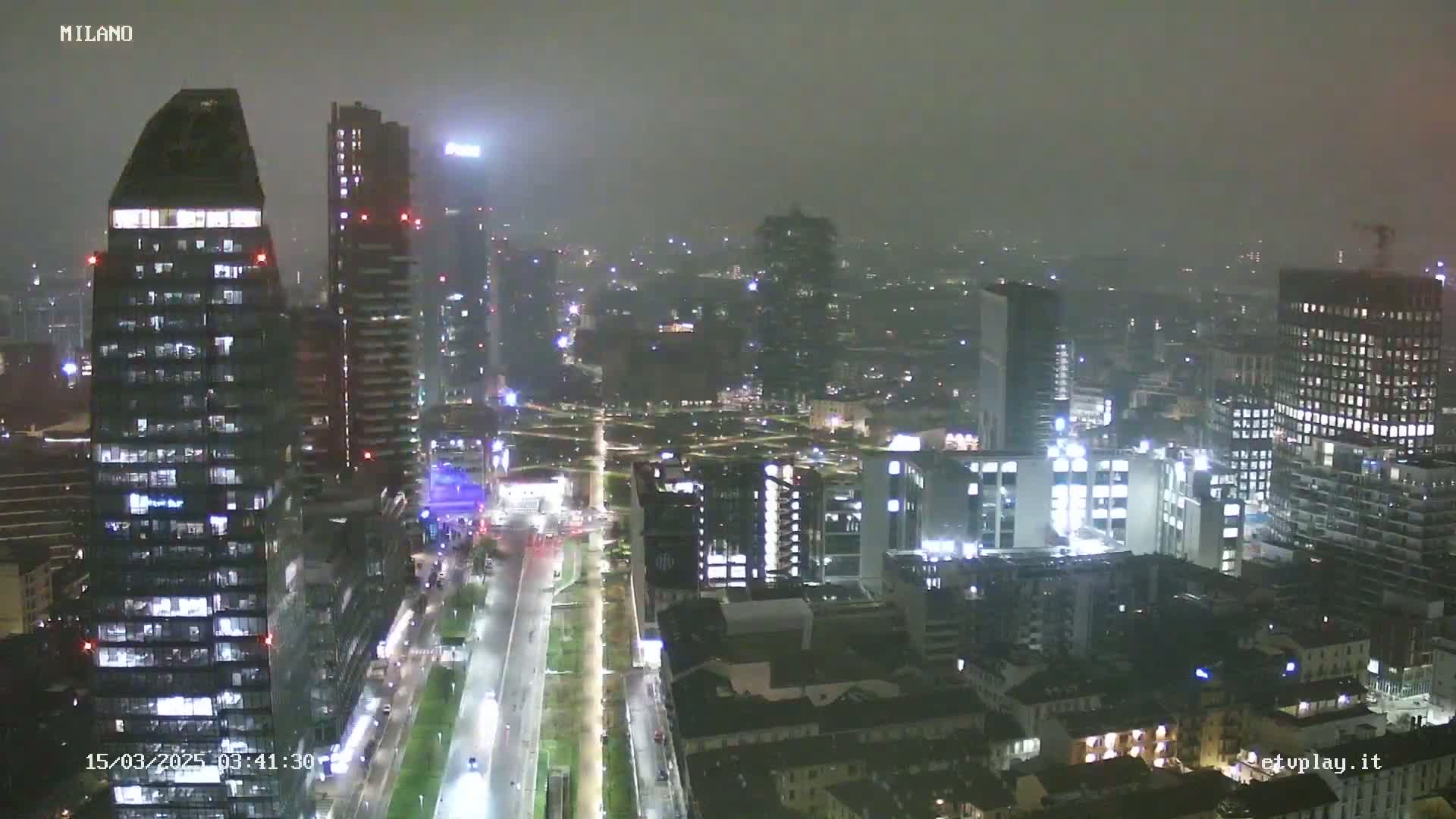 A nighttime aerial view of a city skyline shrouded in fog or mist, showing numerous illuminated buildings and a long, straight road.