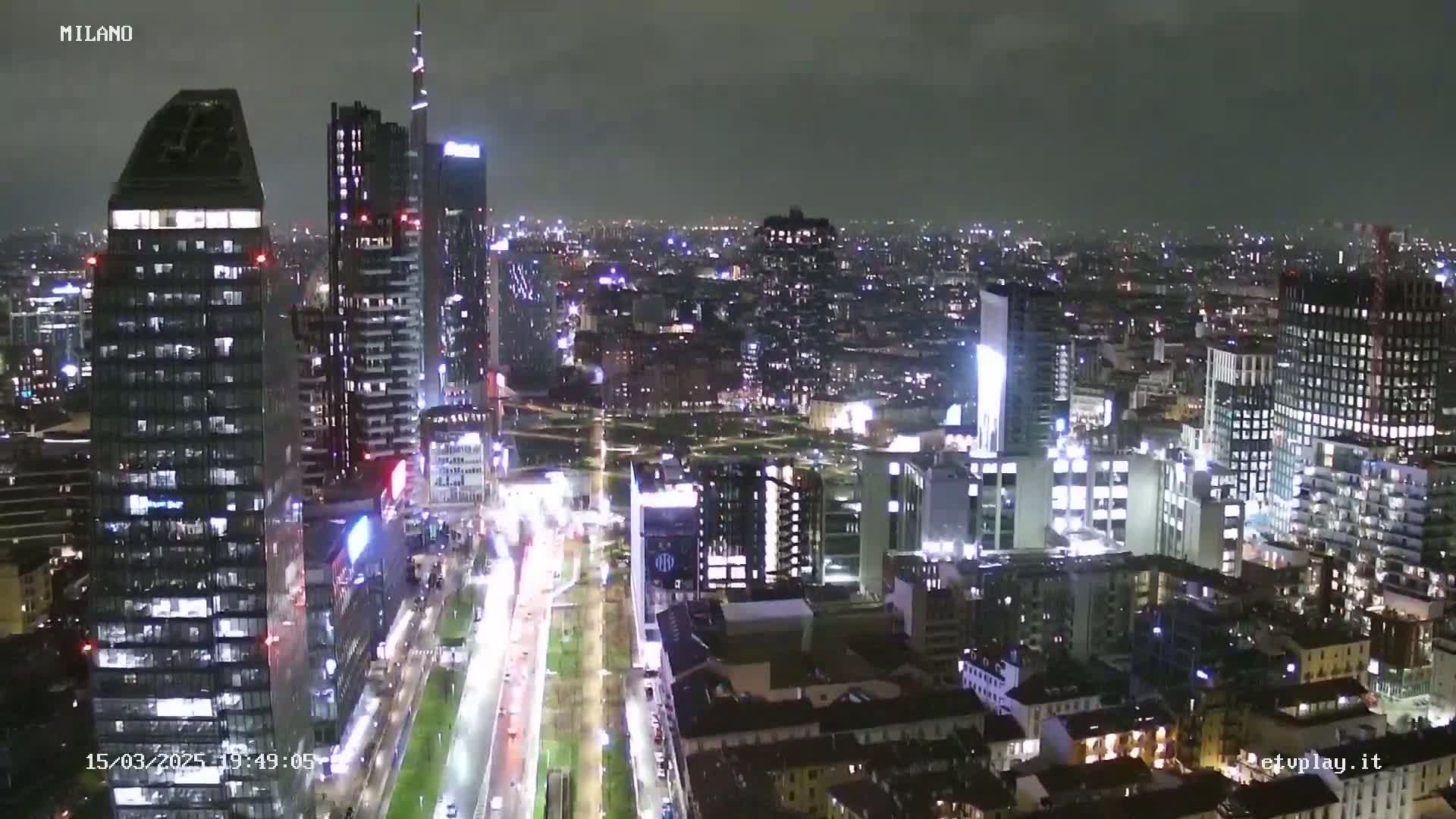 A nighttime view of a city's skyline, showing numerous illuminated skyscrapers and city streets with moving traffic under a dark, cloudy sky.