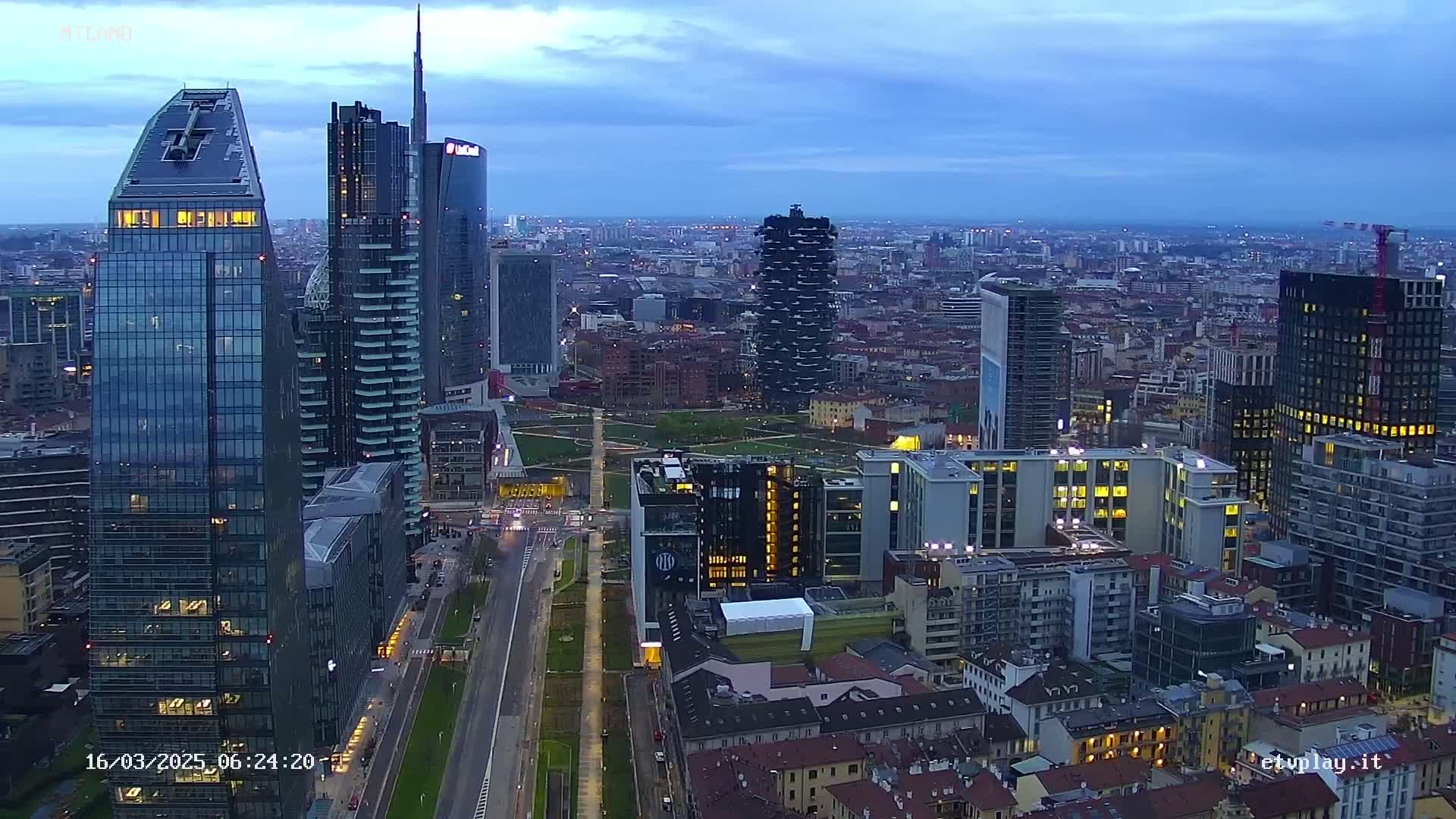 An aerial view of a city skyline at dusk, showing numerous skyscrapers and buildings with some lights on, under a mostly clear sky.