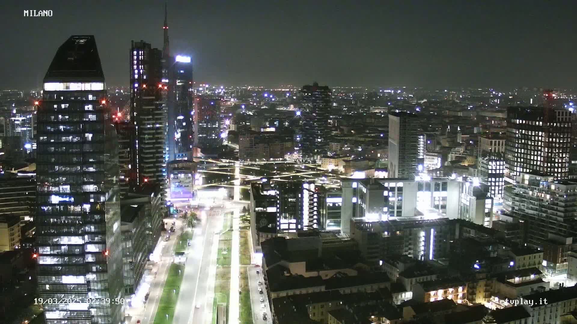 A nighttime aerial view of a city skyline, showing numerous illuminated skyscrapers and buildings stretching into the distance under a clear, dark sky.