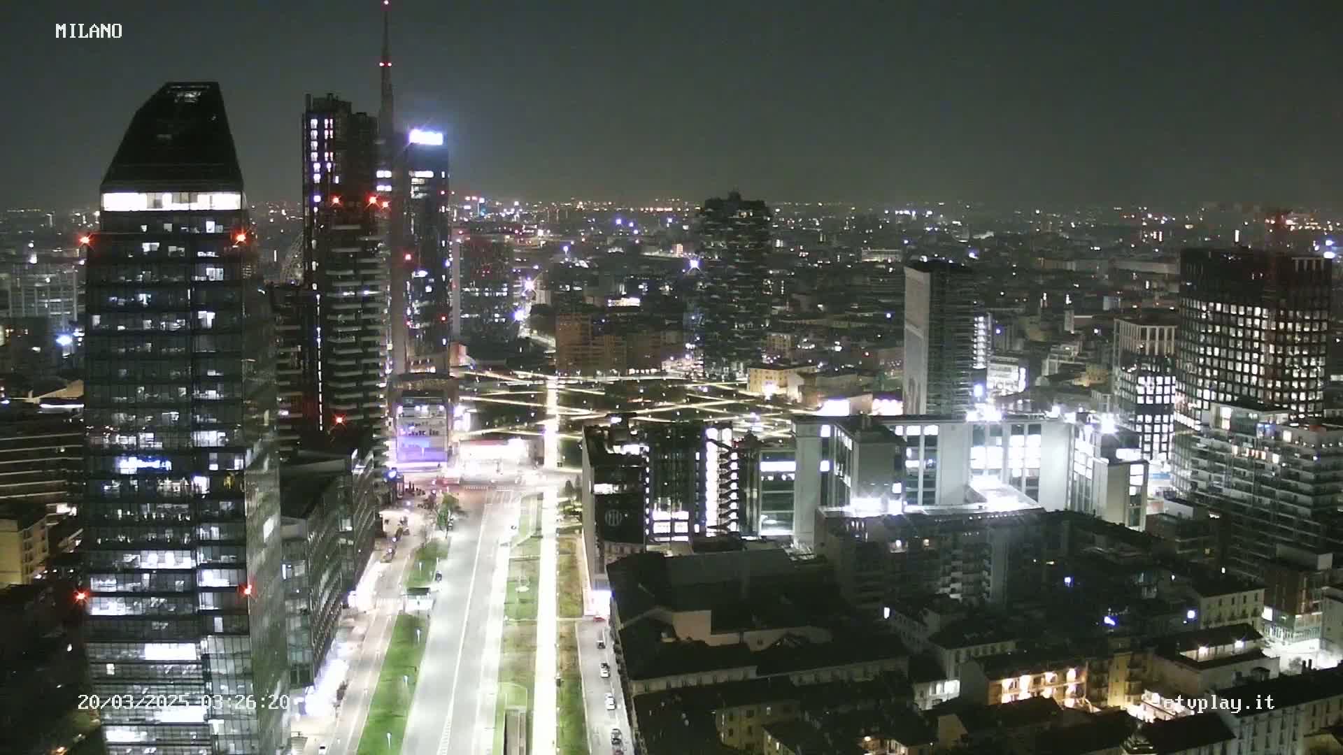 A nighttime aerial view of a city, showing numerous illuminated skyscrapers and buildings extending into the distance under a dark sky.