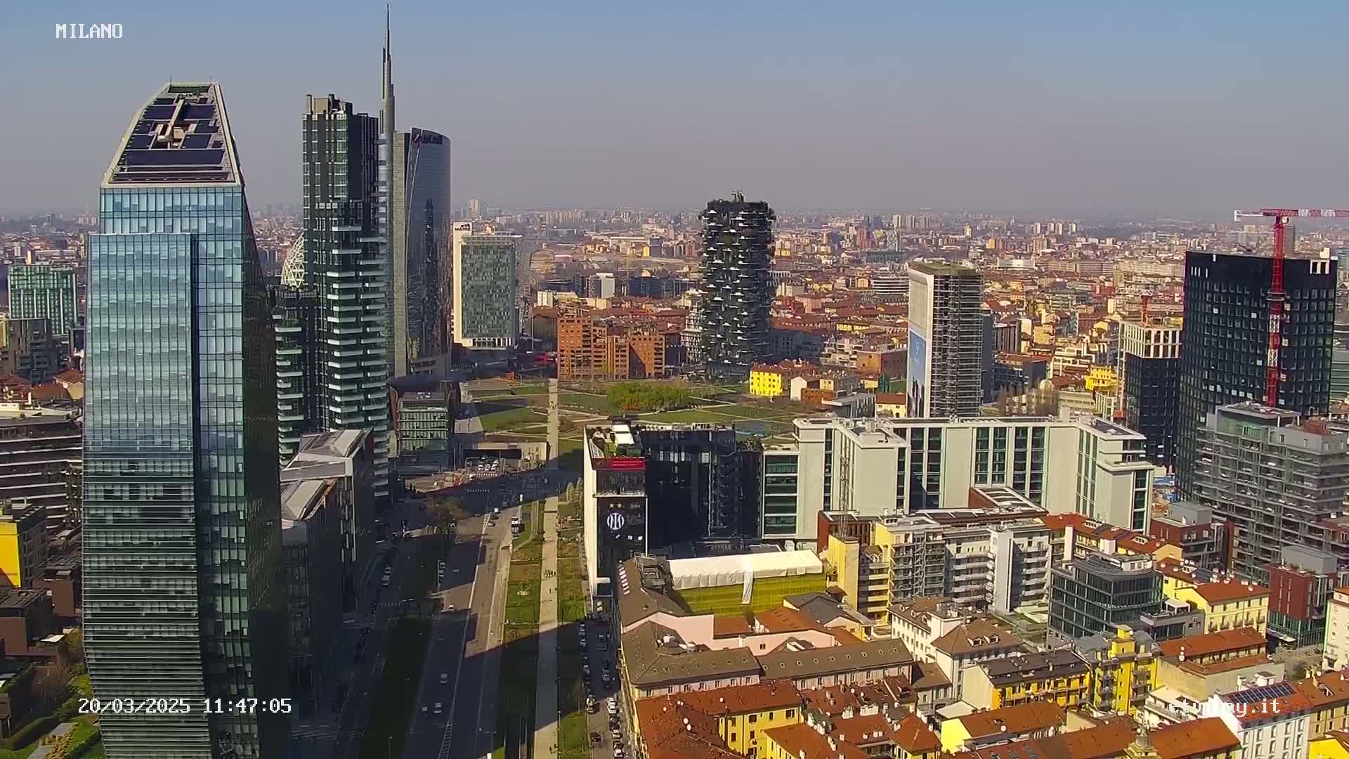 A high-angle, long shot of a dense cityscape on a clear, sunny day, showing many modern skyscrapers and surrounding buildings.