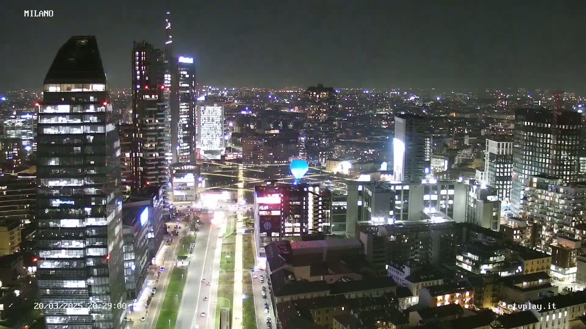 A nighttime aerial view of a city skyline with many illuminated skyscrapers and a road below, under a clear, dark sky.