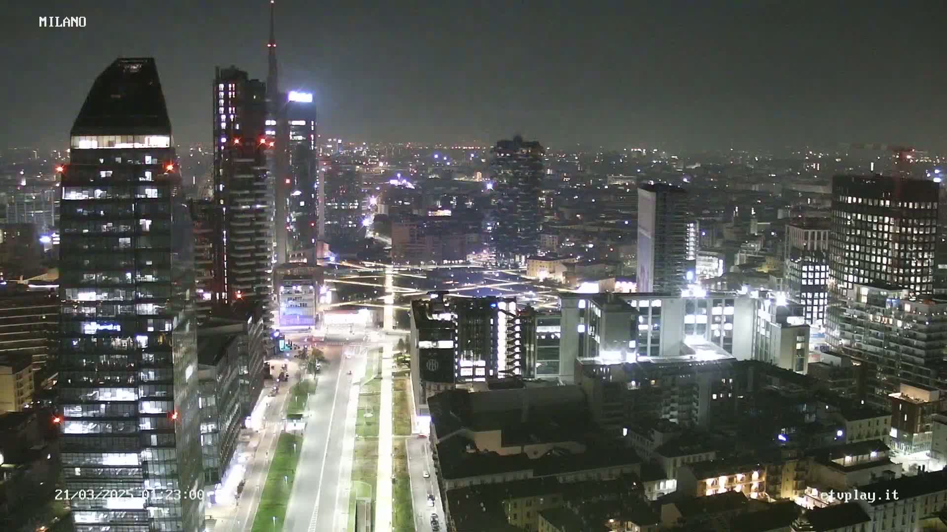 A nighttime aerial view of a city skyline, showing numerous illuminated skyscrapers and buildings, with a long, brightly lit road cutting through the scene.
