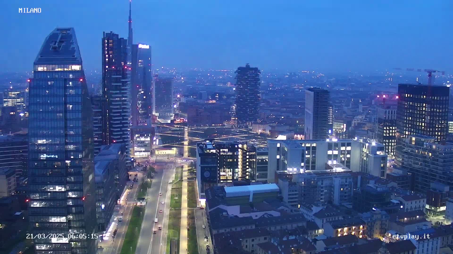 A nighttime aerial view of a city with many skyscrapers and brightly lit streets under a twilight sky.