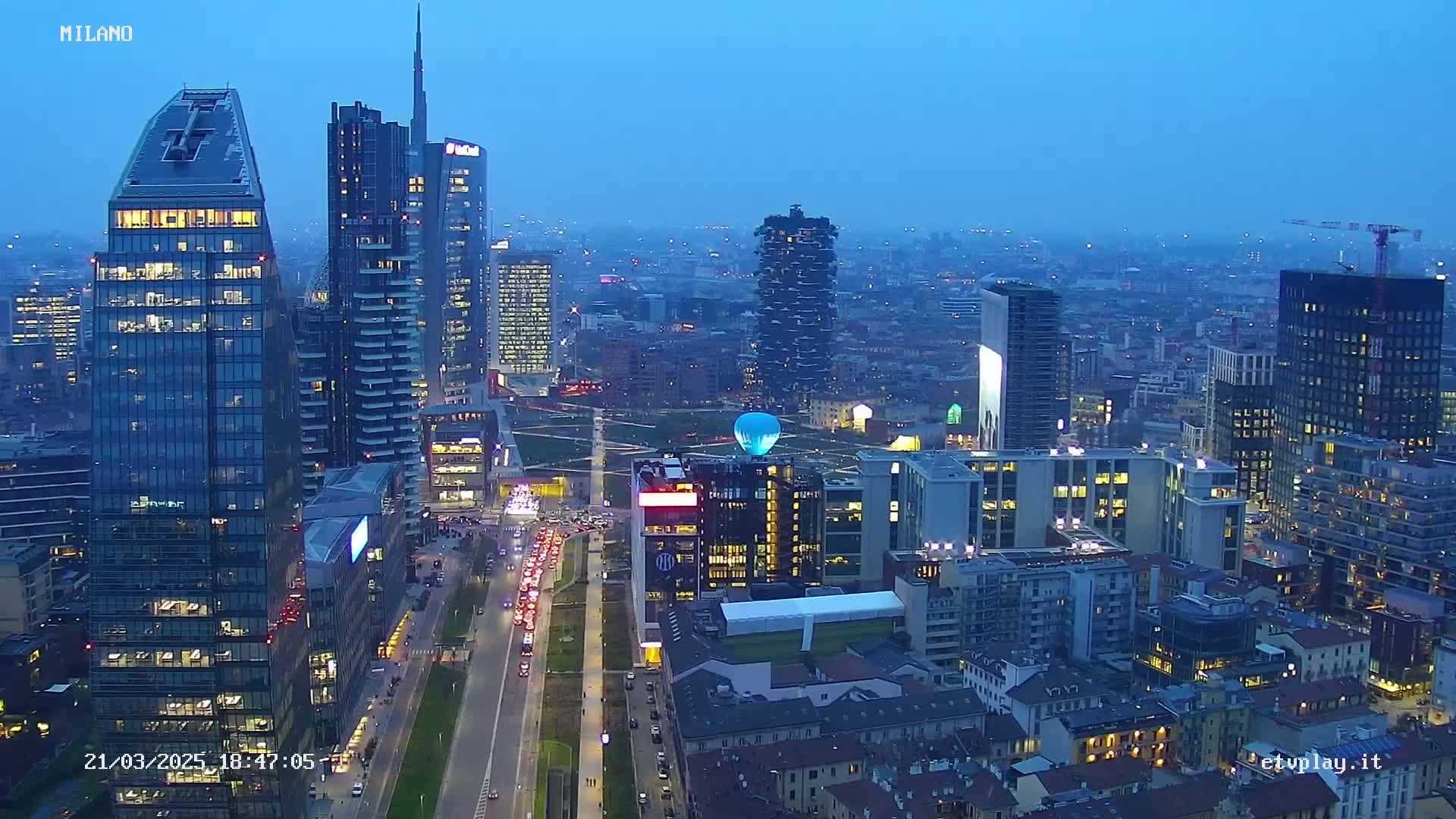 A nighttime aerial view of a city's skyline, showing numerous illuminated skyscrapers and a light-blue hot air balloon, under a twilight sky.