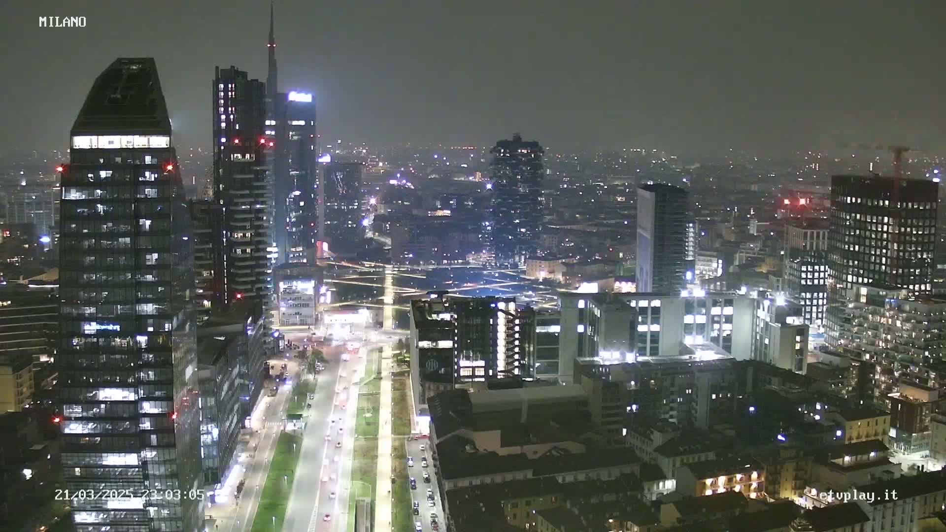 A nighttime aerial view of a city, showing numerous illuminated skyscrapers and city streets under hazy conditions.