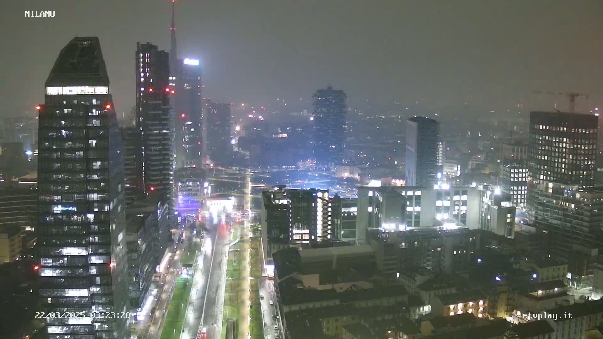 A nighttime view of a city skyline with many tall buildings under hazy, rainy conditions.