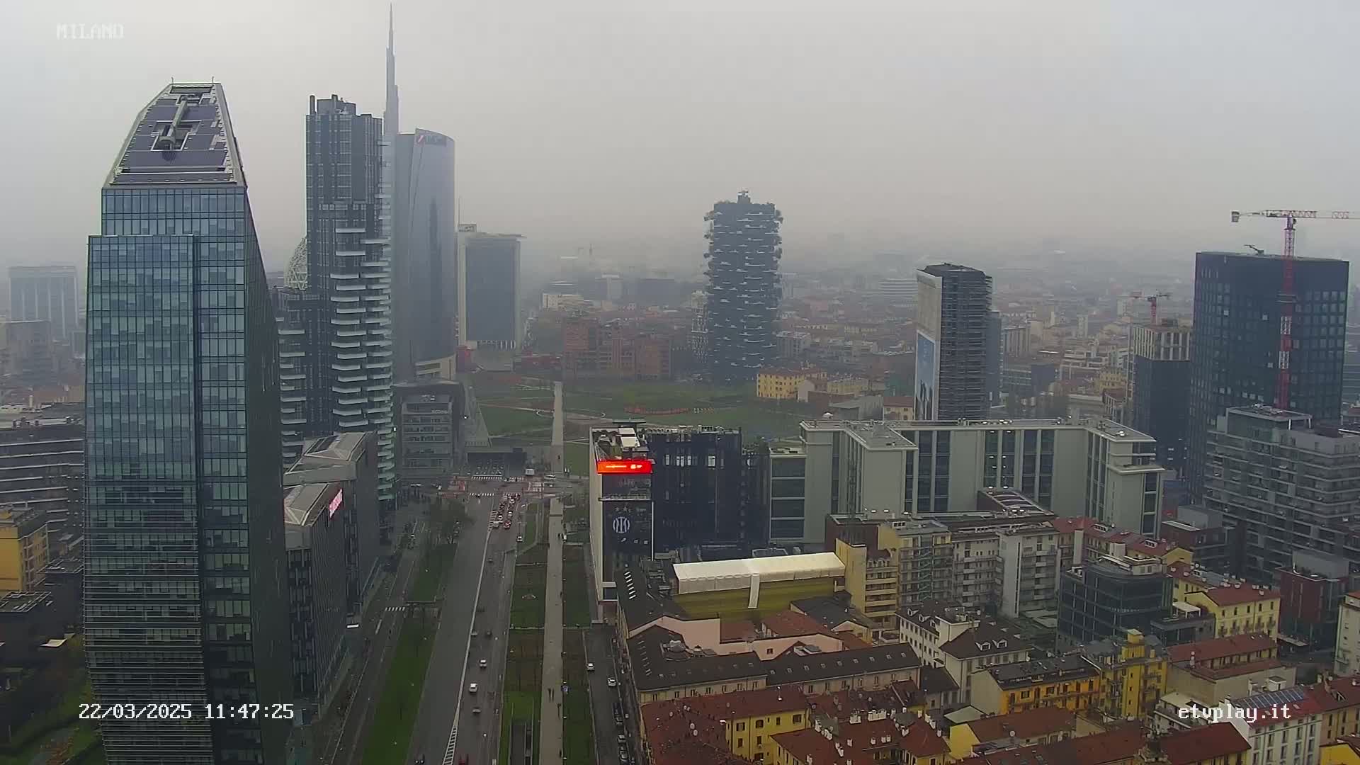 A hazy, overcast cityscape view shows numerous tall buildings and a wide street, with some construction visible.