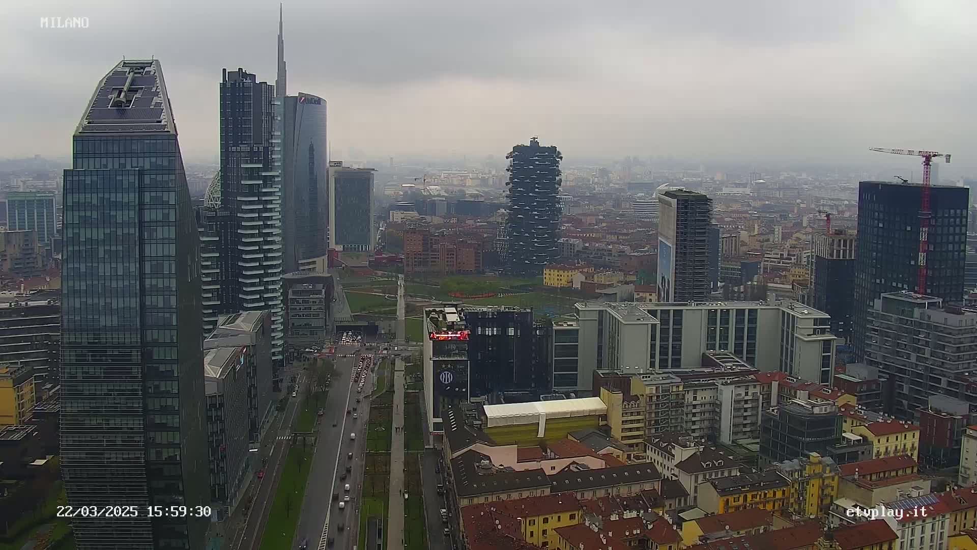 An aerial view of a city skyline on an overcast day, showing numerous skyscrapers and high-rise buildings, with some construction cranes visible.
