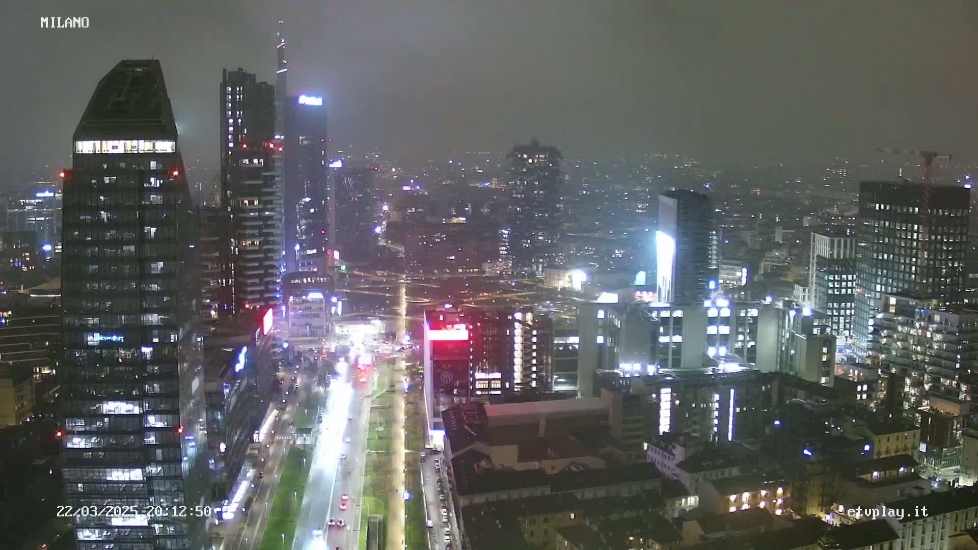 A nighttime view of a city skyline shrouded in fog or mist, showing numerous illuminated skyscrapers and a long, brightly lit street below.