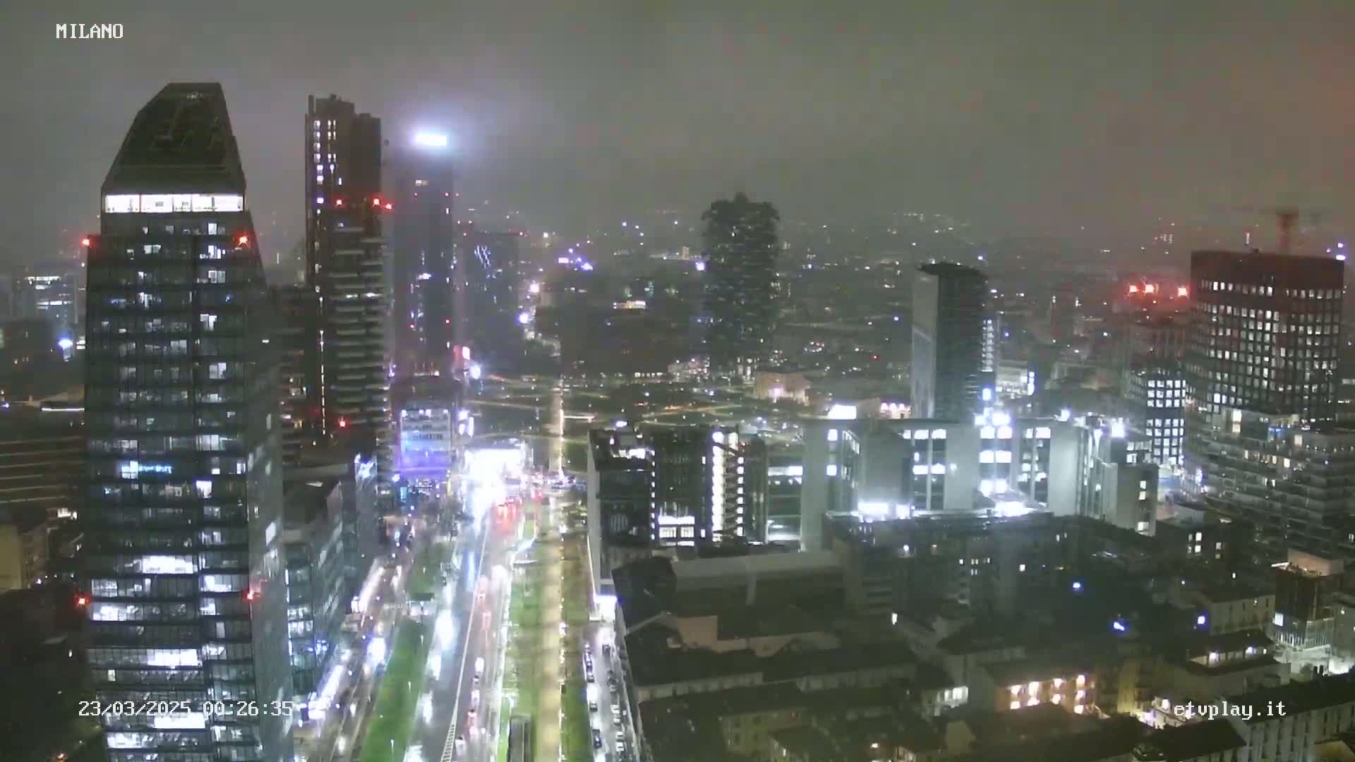 A nighttime view of a city skyline with many tall buildings illuminated, under hazy or foggy conditions.