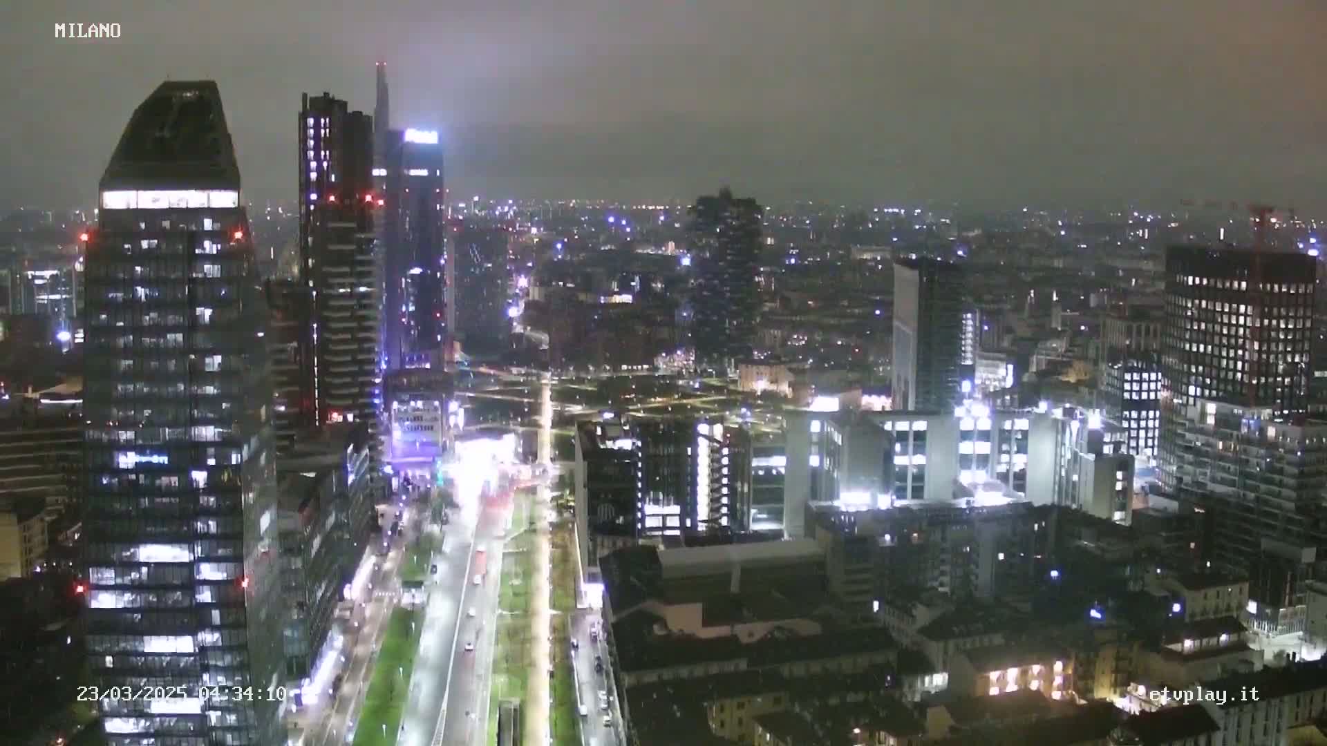 A nighttime aerial view of a city with many illuminated skyscrapers and buildings, under a hazy sky.