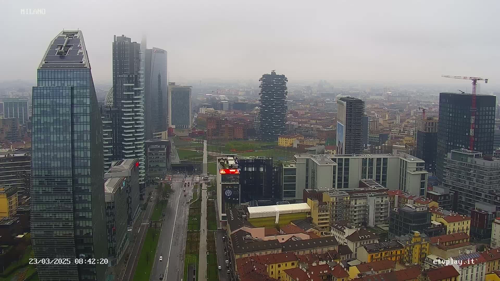 A high-angle, long shot of a city skyline on an overcast day shows numerous skyscrapers and other buildings, with a wide street and park visible below.