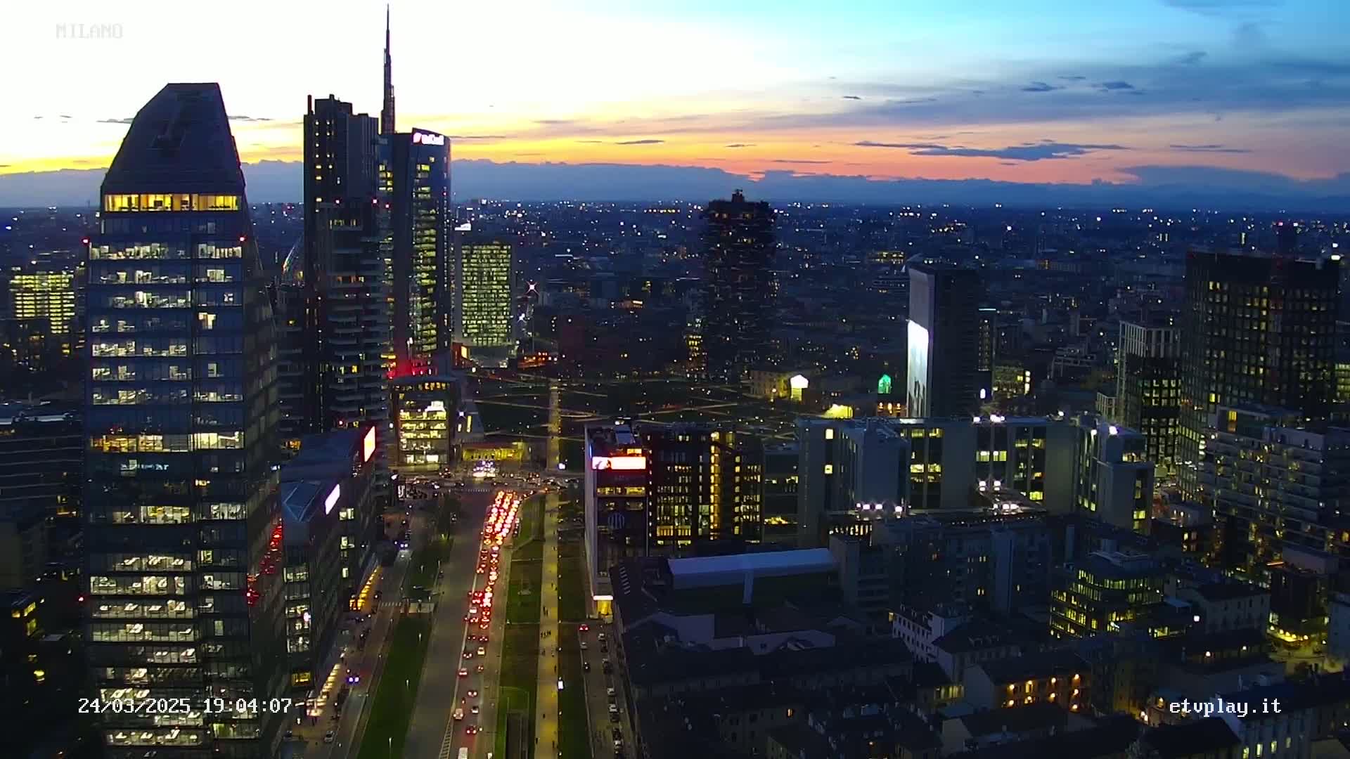 A city skyline at dusk, showing numerous illuminated skyscrapers and a busy road with traffic, under a colorful sunset sky.