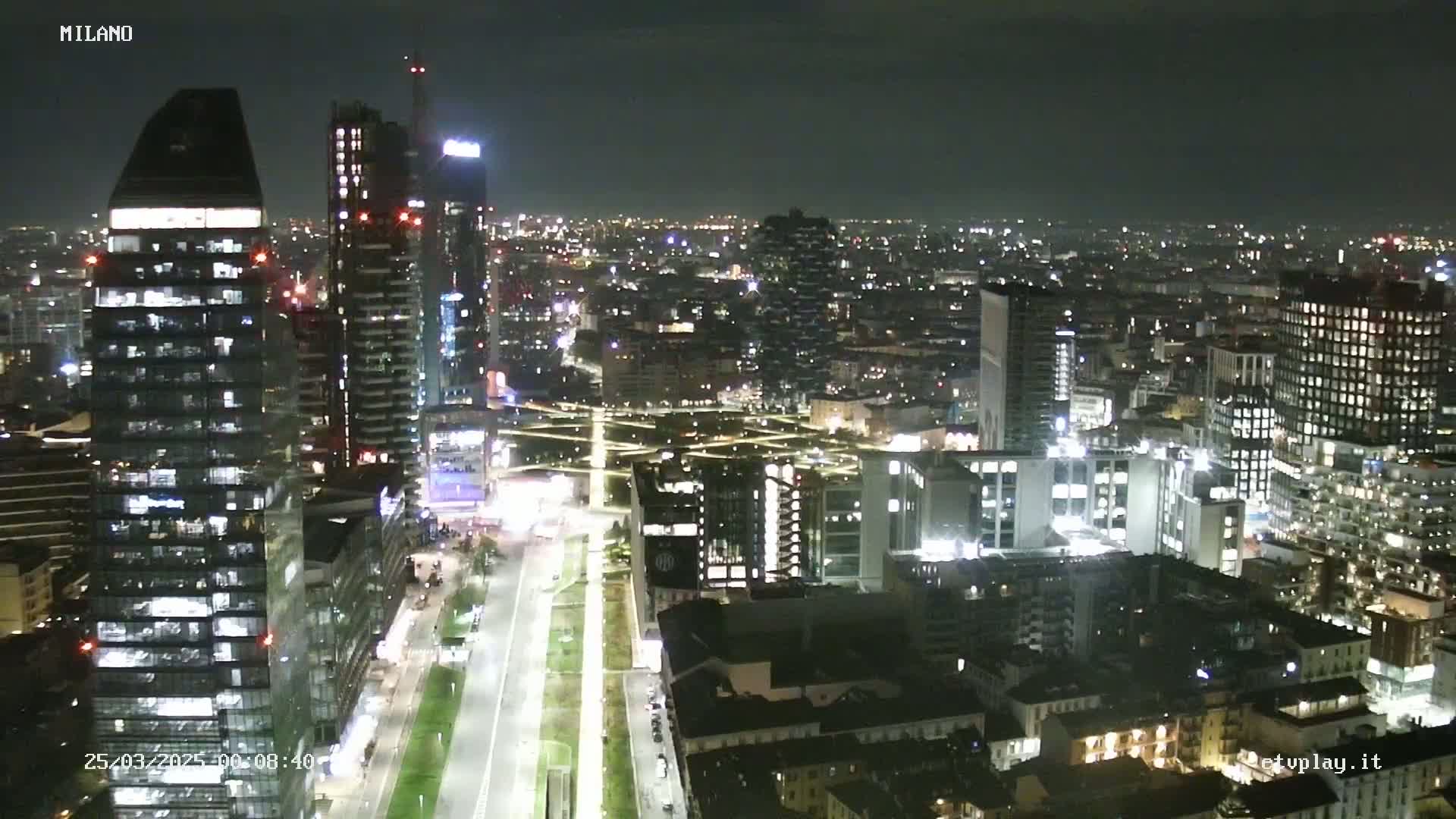 A nighttime aerial view of a city illuminated by lights, showing many tall buildings and roads.
