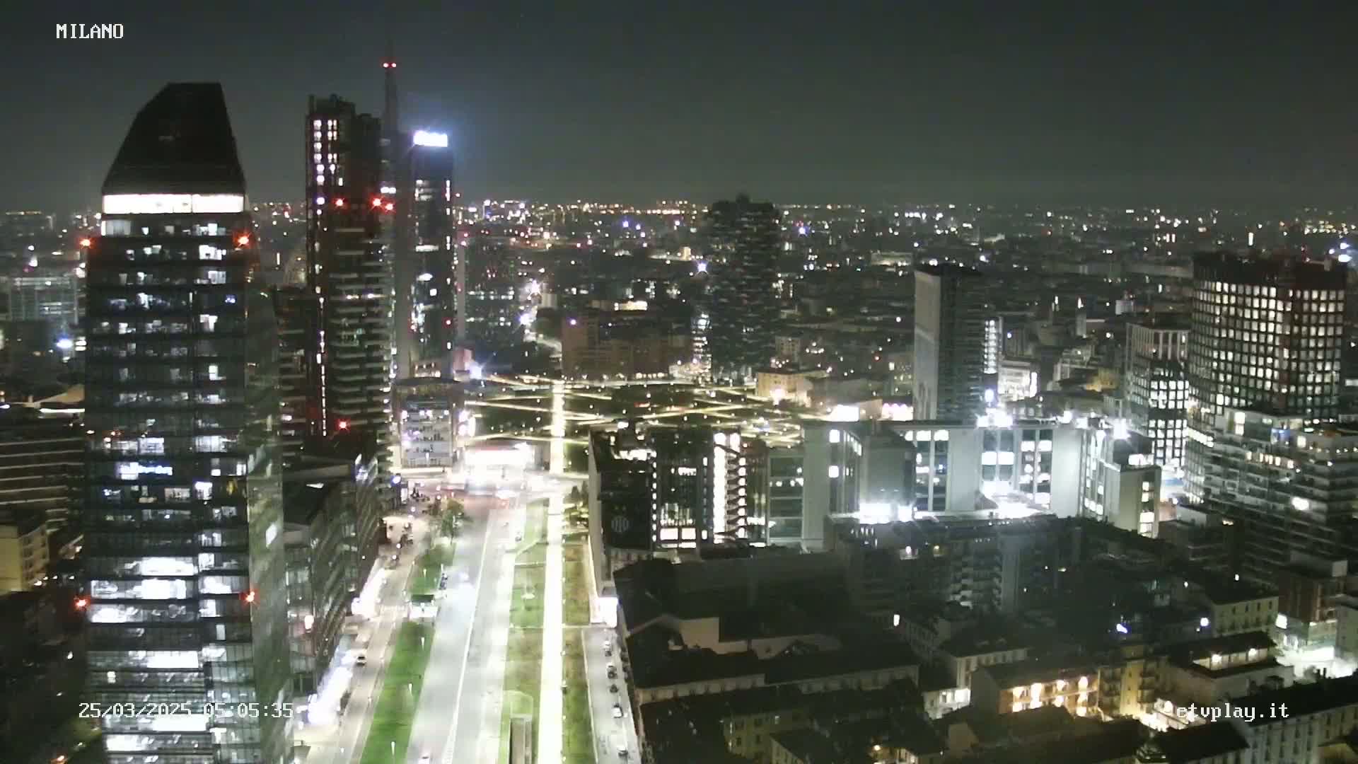 A nighttime aerial view of a city's skyline, showing brightly lit skyscrapers and streets with cars, under a clear, dark sky.