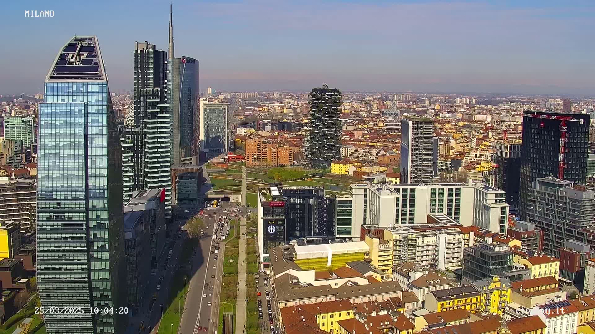 A sunny aerial view of a city with numerous skyscrapers and a sprawling cityscape extending to the horizon.