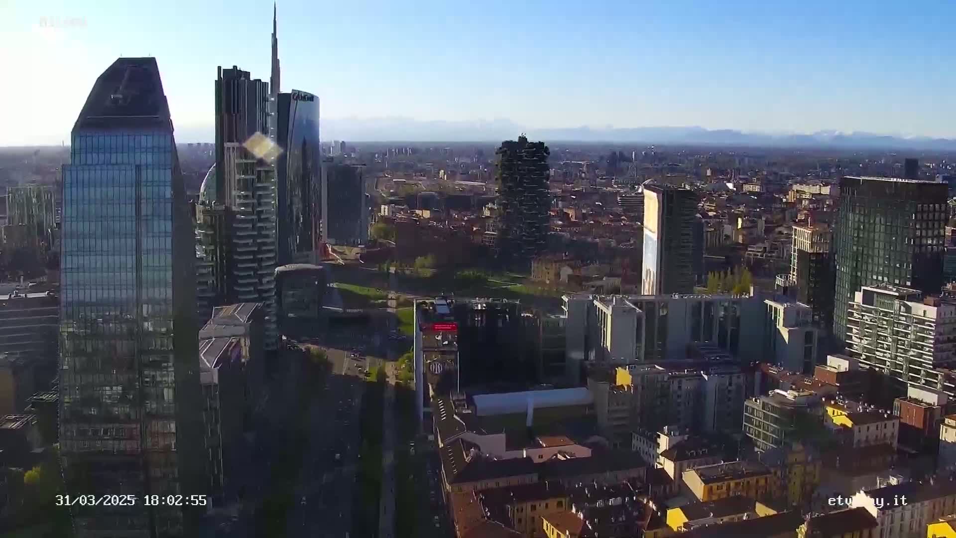 A high-angle, long shot of a city skyline on a clear, sunny day, showing numerous skyscrapers and surrounding buildings, with mountains visible in the distance.