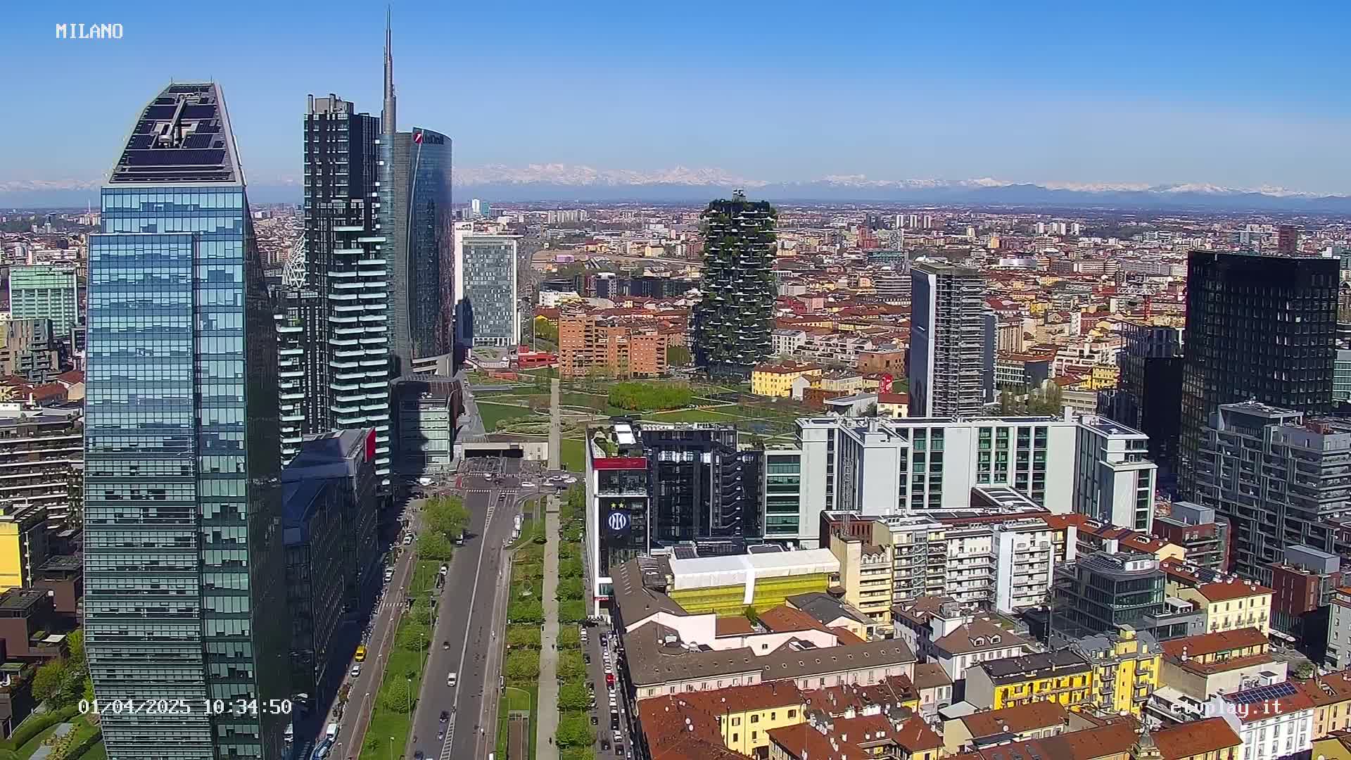 A high-angle, long shot of a dense city, featuring numerous skyscrapers and other buildings, under a clear blue sky with snow-capped mountains visible in the distance.