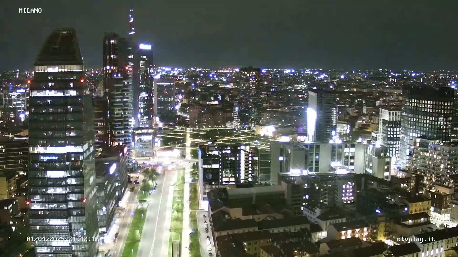 A nighttime aerial view of a city skyline, showing brightly lit skyscrapers and streets, under a clear sky.