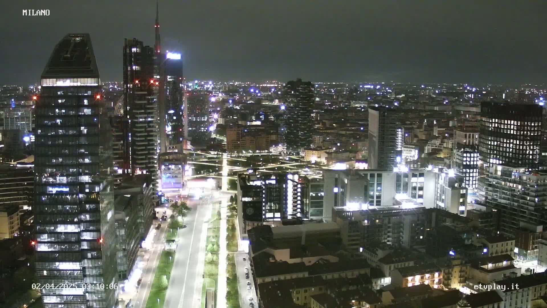 A nighttime aerial view of a densely populated city with numerous illuminated skyscrapers and buildings, extending into a dimly lit horizon under a dark sky.