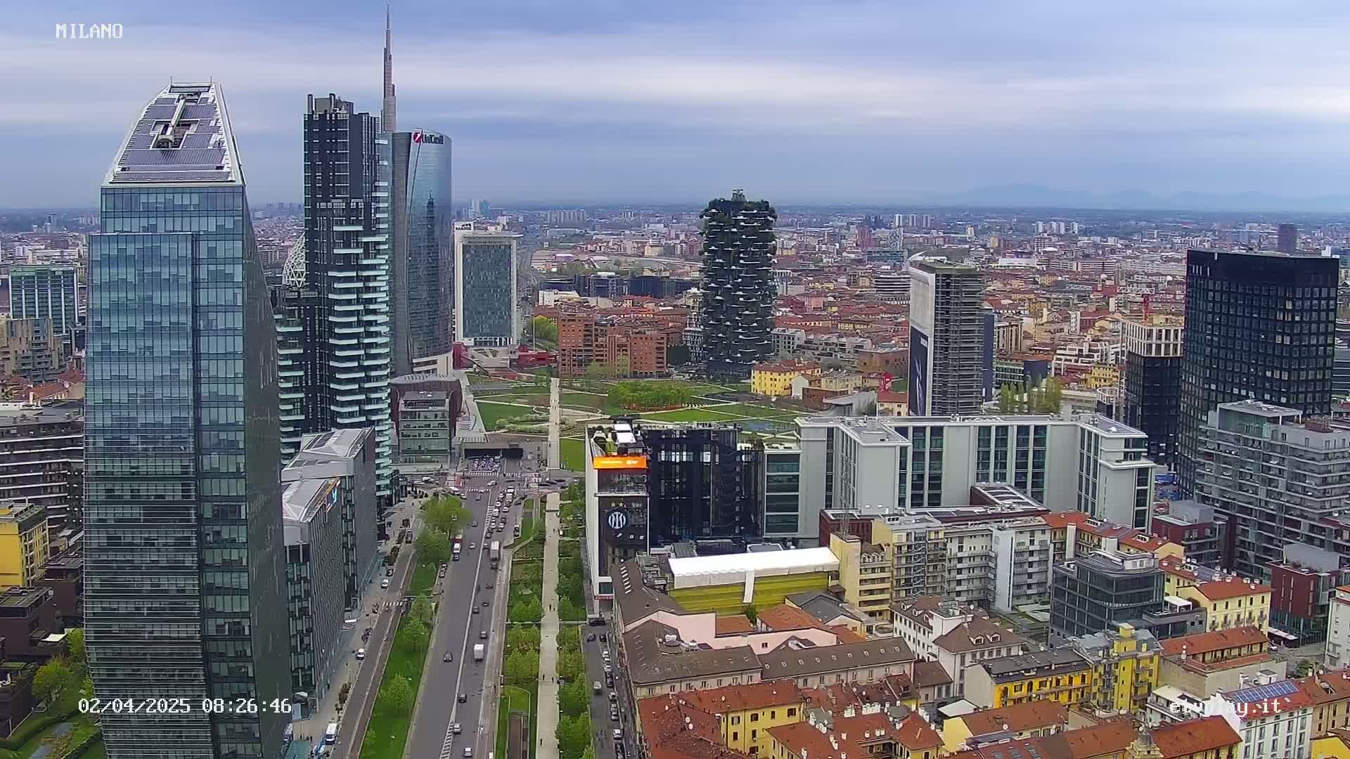 An aerial view of a city skyline on a partly cloudy day shows many modern skyscrapers and a mix of older buildings, with a wide road and green space visible between clusters of buildings.