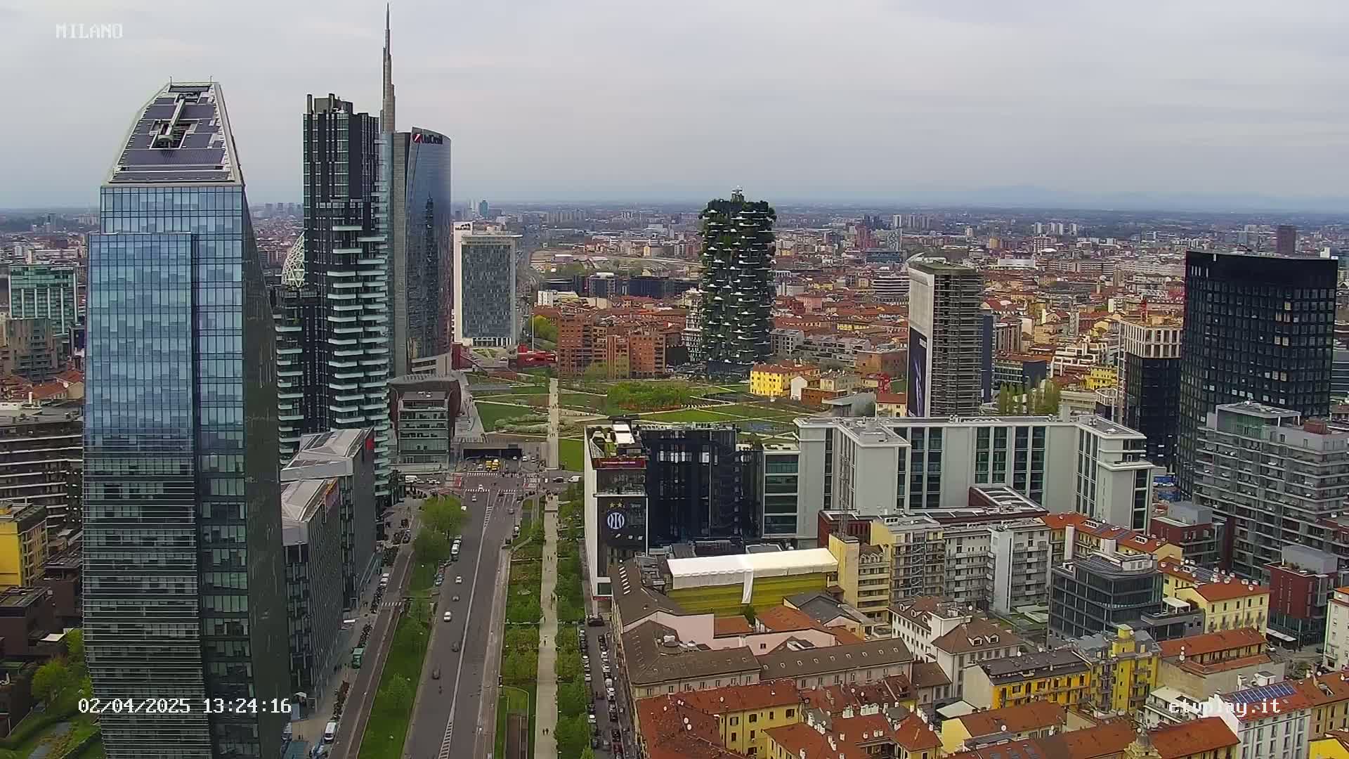 An aerial view shows a cityscape of numerous skyscrapers and high-rise buildings, with a park visible in the center, under a cloudy sky.
