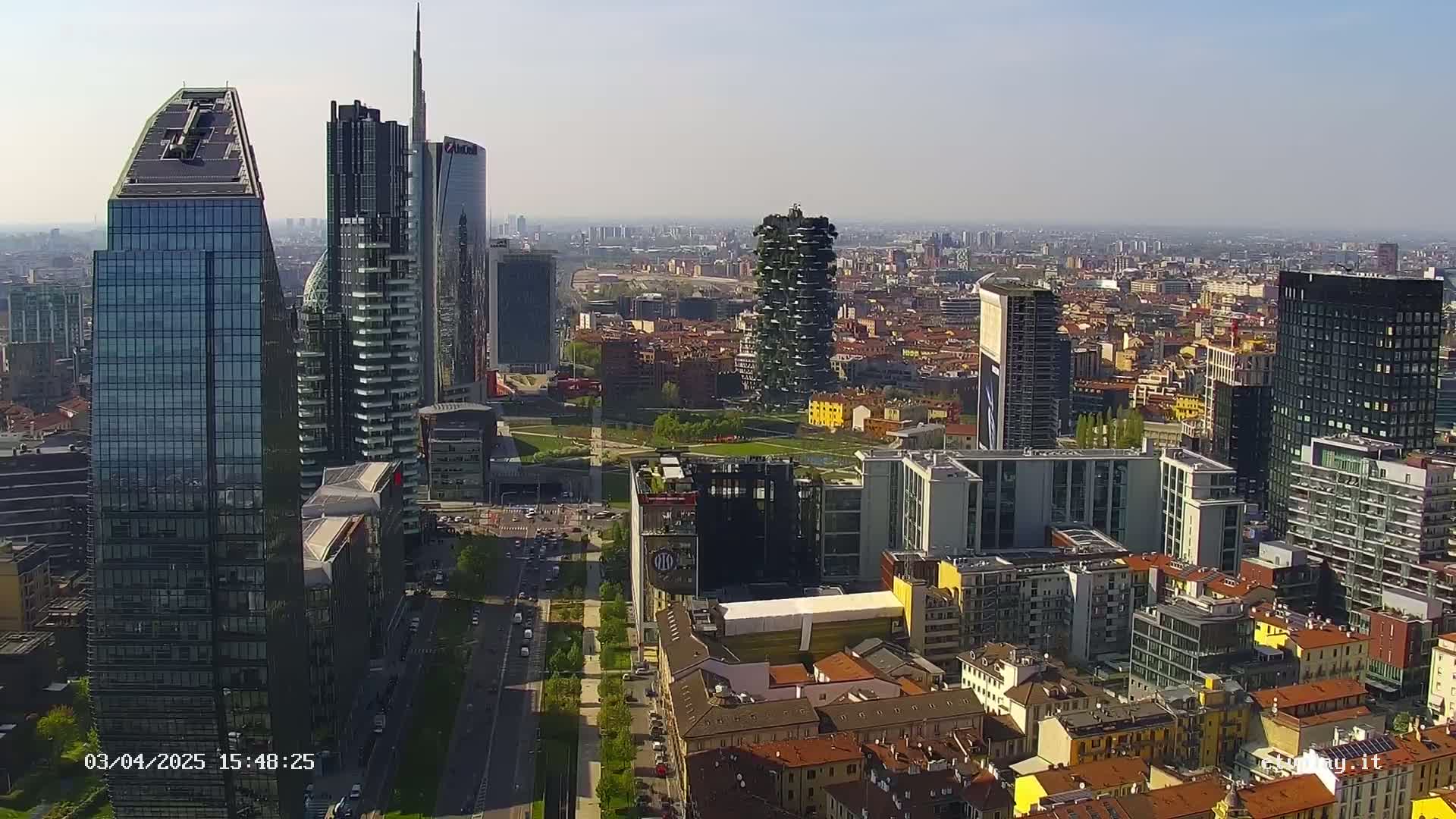 A high-angle, long shot shows a cityscape of numerous skyscrapers and low-rise buildings under a clear, sunny sky.