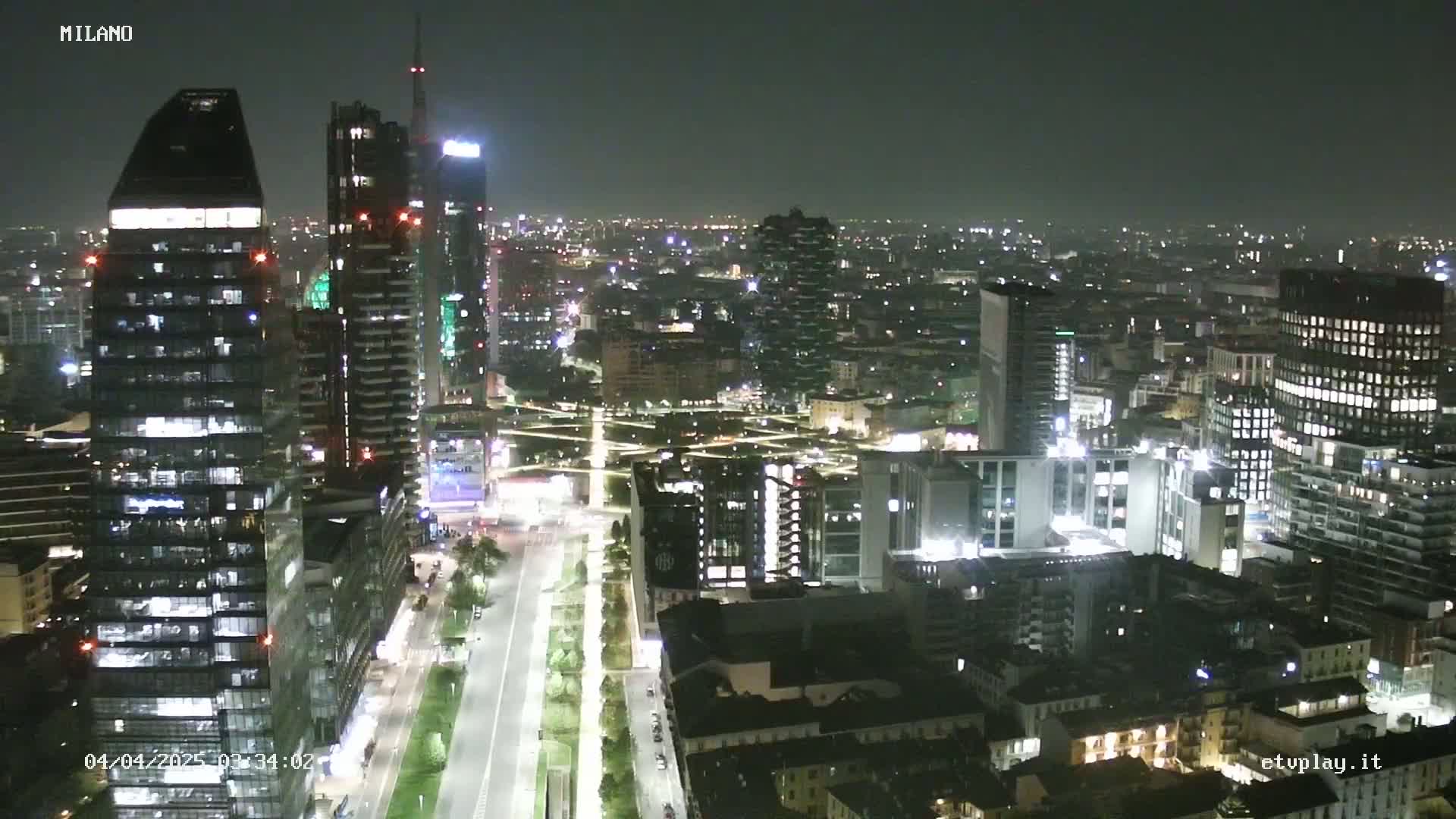 A nighttime aerial view of a city skyline with numerous illuminated buildings and a long, tree-lined street.