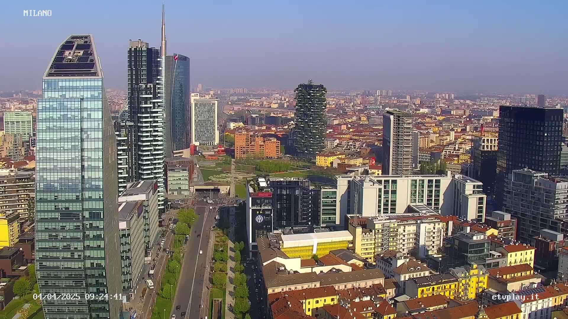 A high-angle, long shot of a modern city skyline on a clear, sunny day, featuring numerous skyscrapers and surrounding buildings.