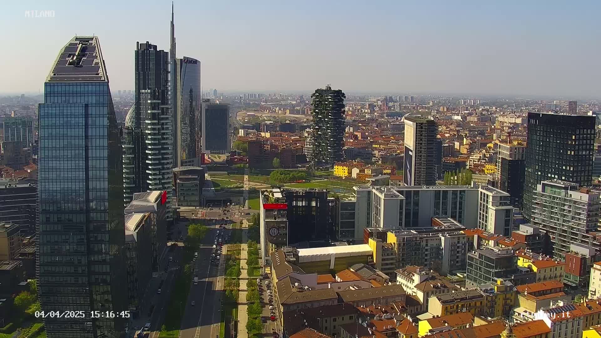 A high-angle, long shot of a city skyline on a clear, sunny day, showing a mix of modern skyscrapers and older, lower buildings.