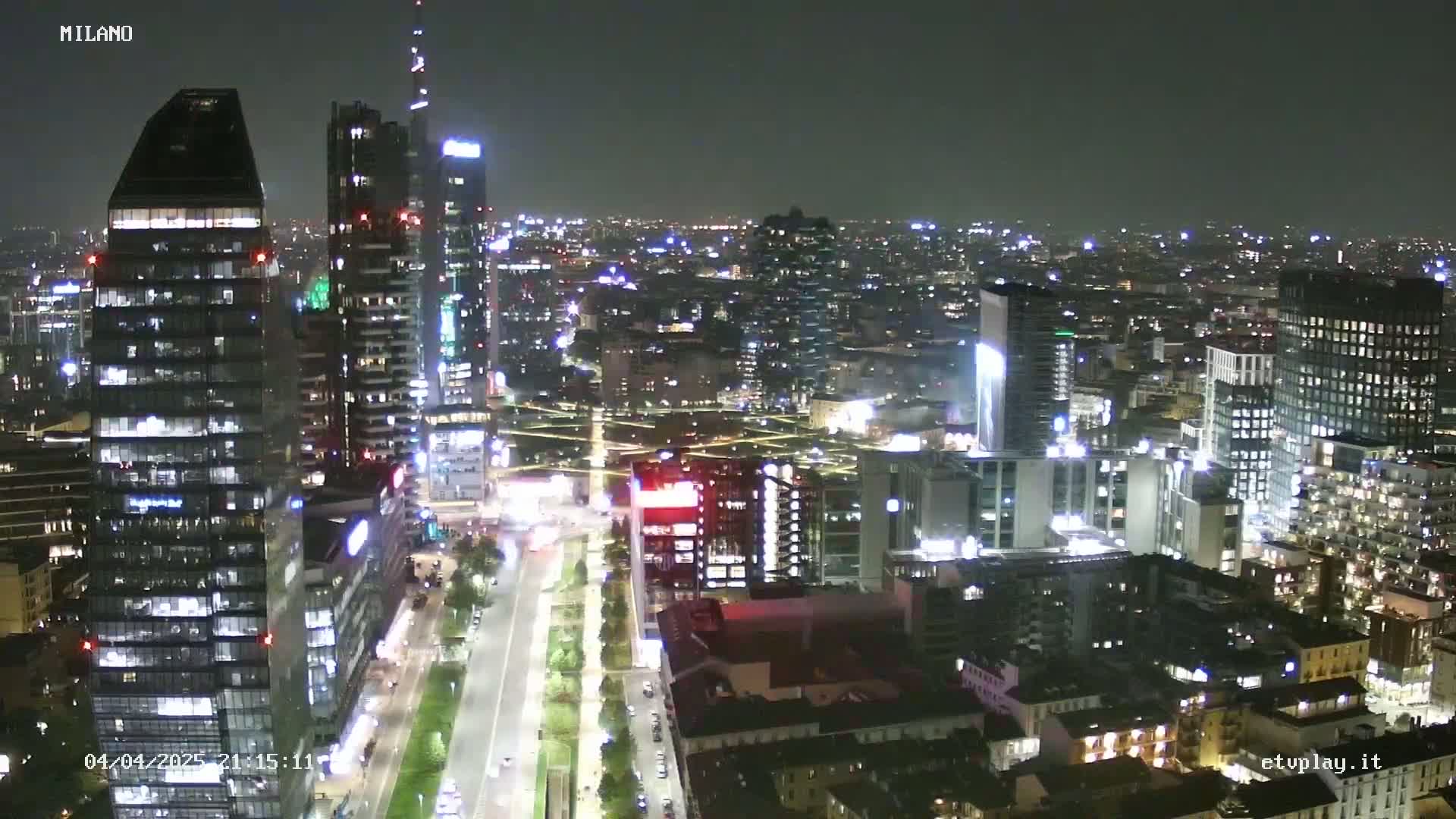 A nighttime aerial view of a city with numerous illuminated skyscrapers and buildings, showing a well-lit street below.
