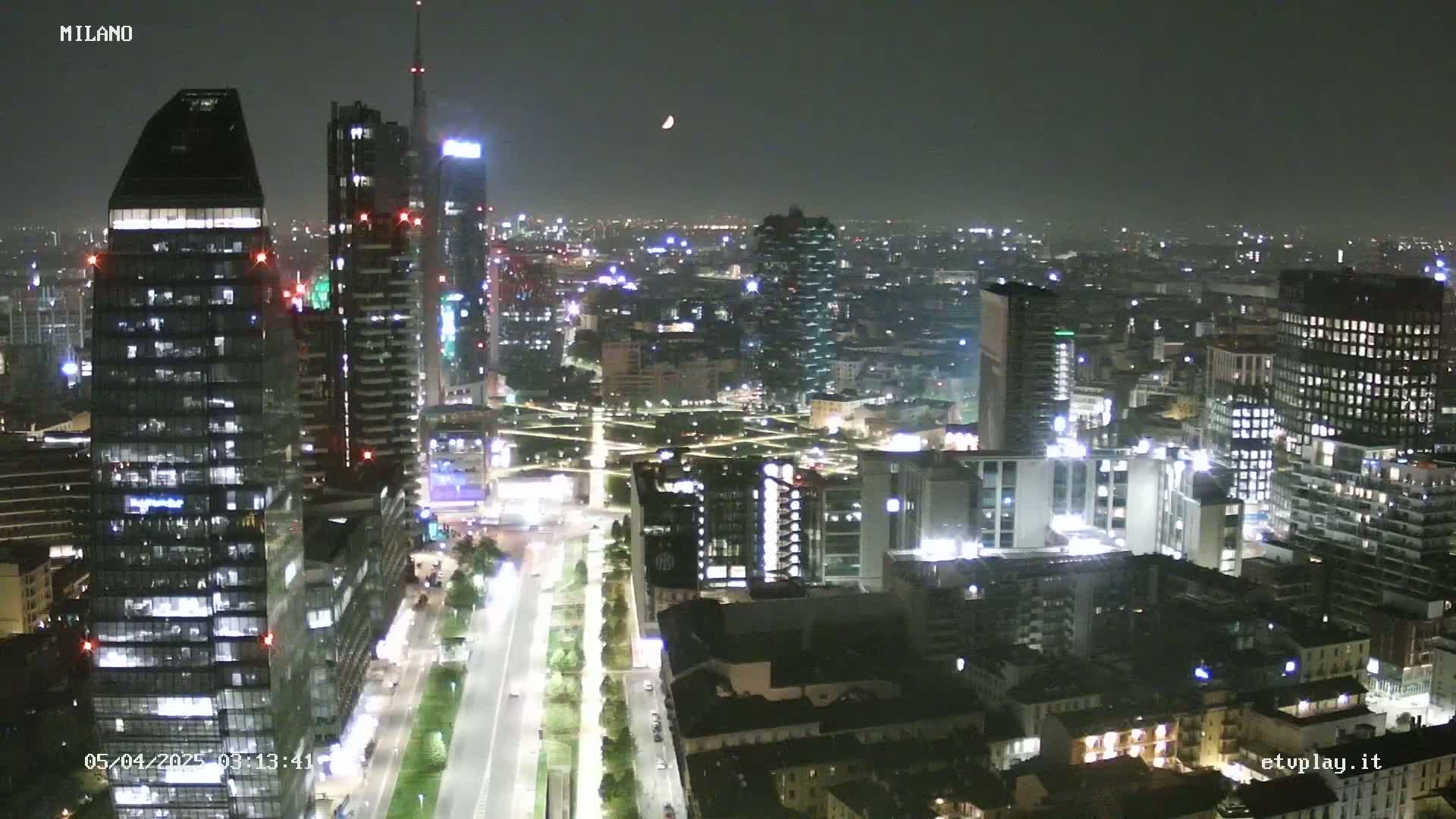 A nighttime aerial view of a city skyline with many lit-up buildings and a crescent moon visible in the dark sky.