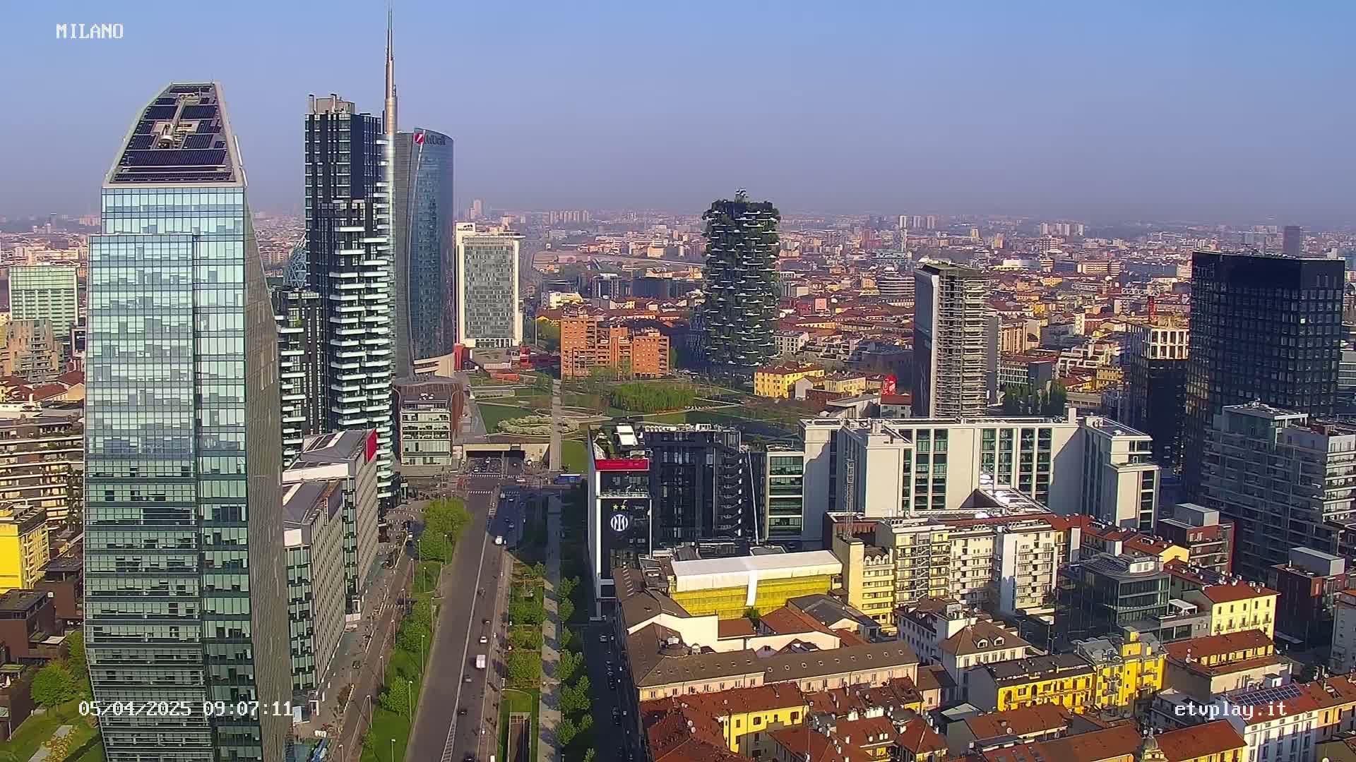 A high-angle, long shot of a dense cityscape on a clear, sunny day, showcasing numerous skyscrapers and a mix of modern and older buildings.