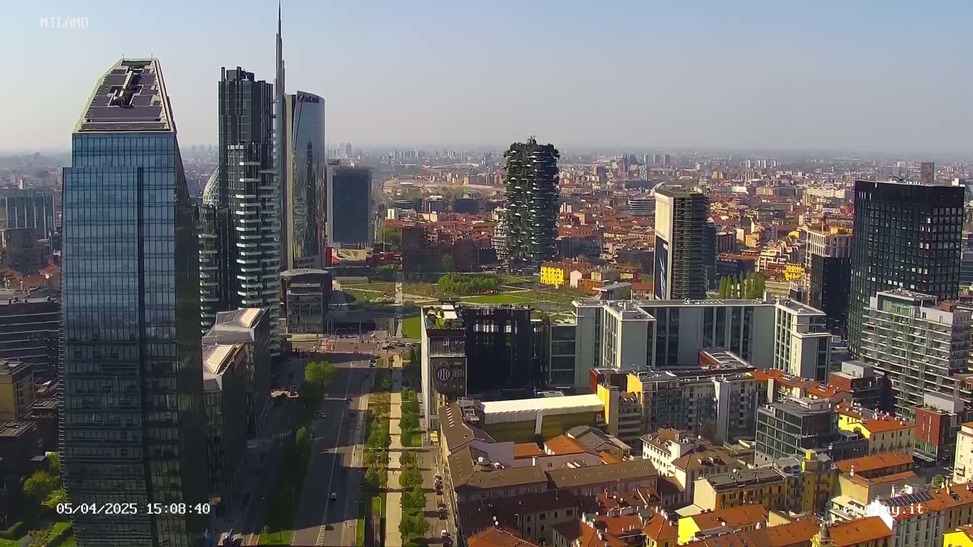 A high-angle, long shot of a city skyline on a clear, sunny day, showing a mix of modern skyscrapers and lower buildings.