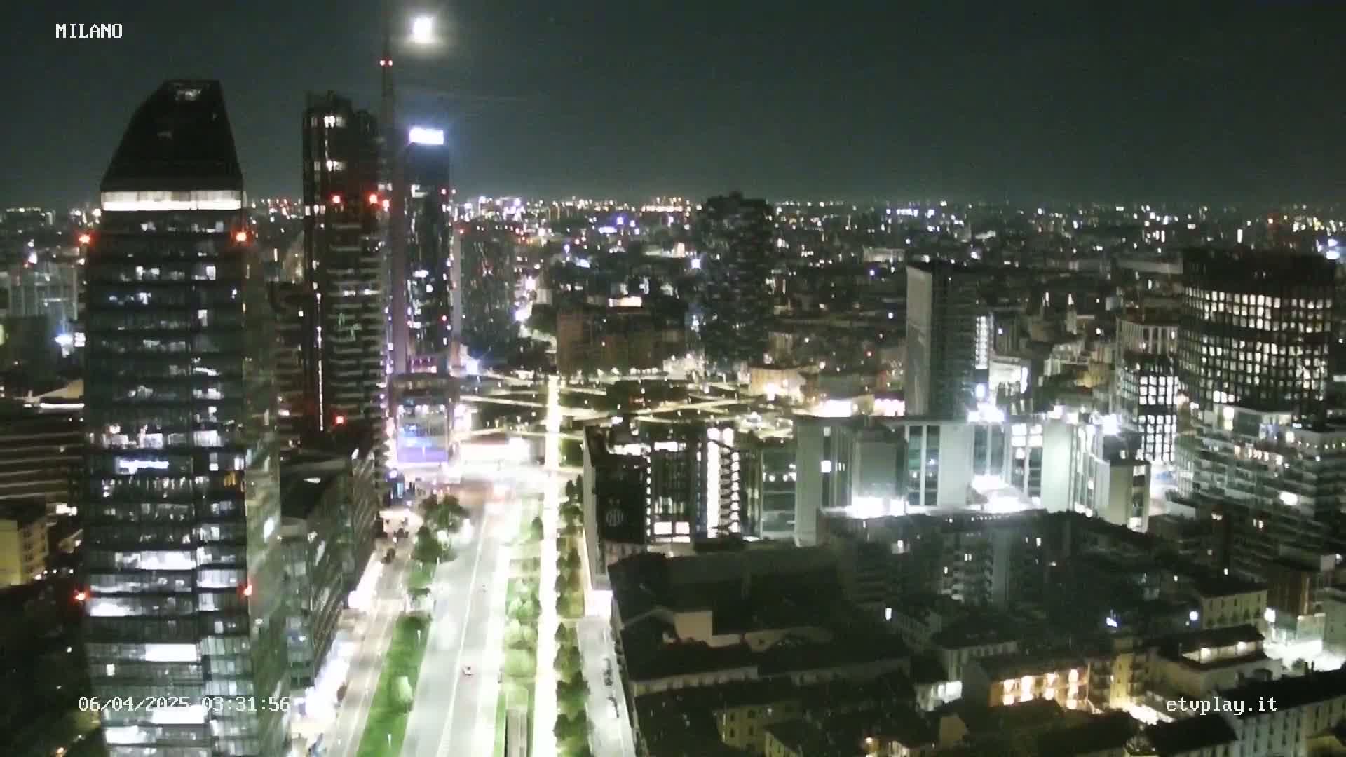 A nighttime aerial view of a city shows numerous illuminated buildings, streets, and trees under a clear sky.