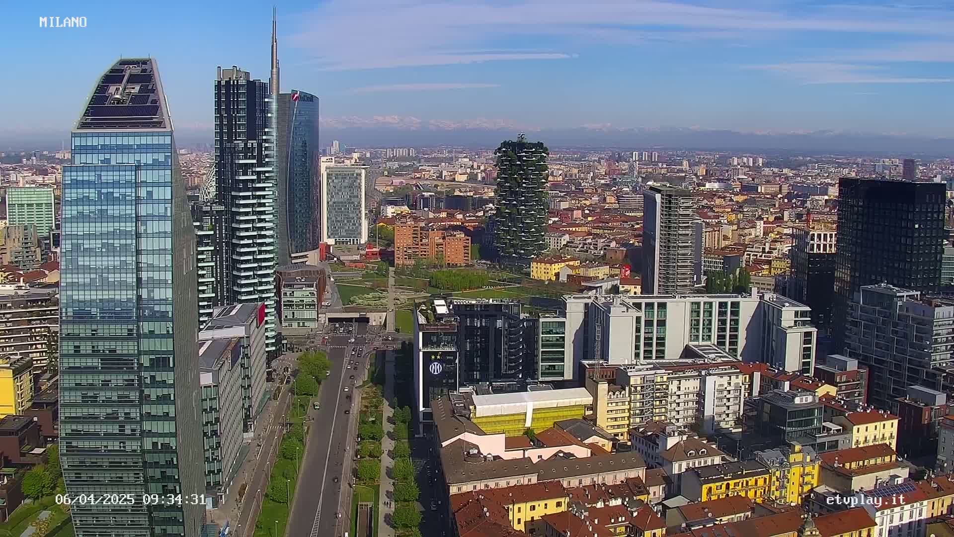 A high-angle, sunny view of a city skyline featuring numerous skyscrapers and a sprawling cityscape, with mountains visible in the distance.