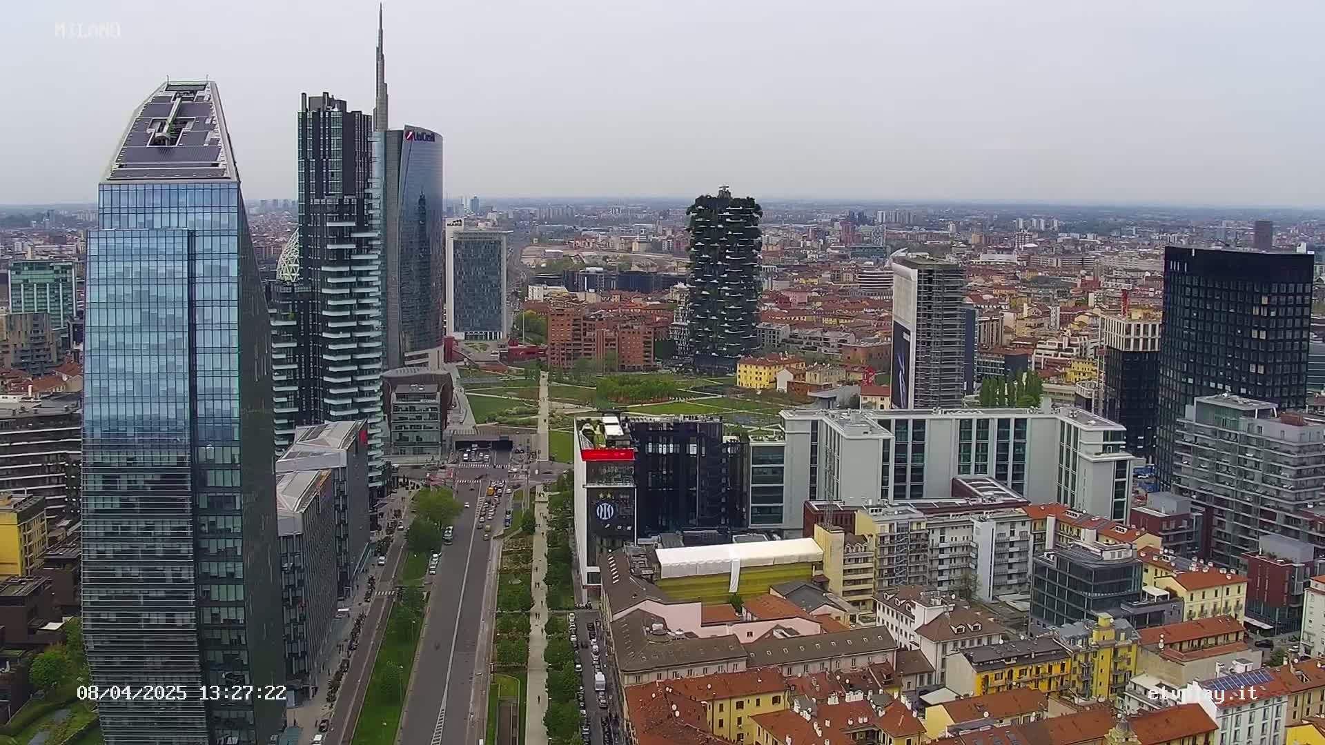 An aerial view of a city with numerous skyscrapers under an overcast sky, showing a wide street and surrounding buildings.
