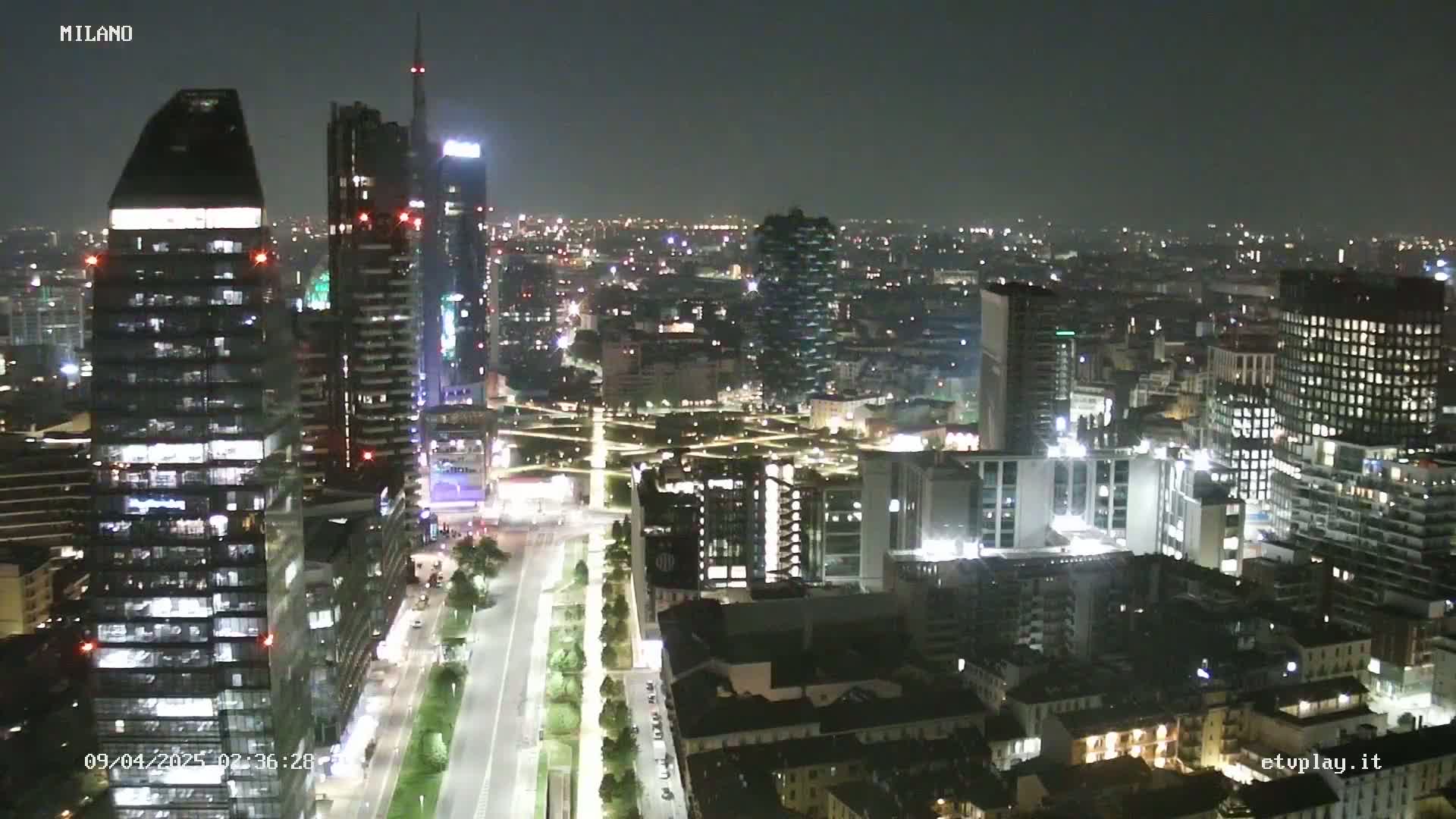 A nighttime aerial view of a city with numerous illuminated skyscrapers and buildings, extending to a darker, more distant cityscape under a clear, dark sky.
