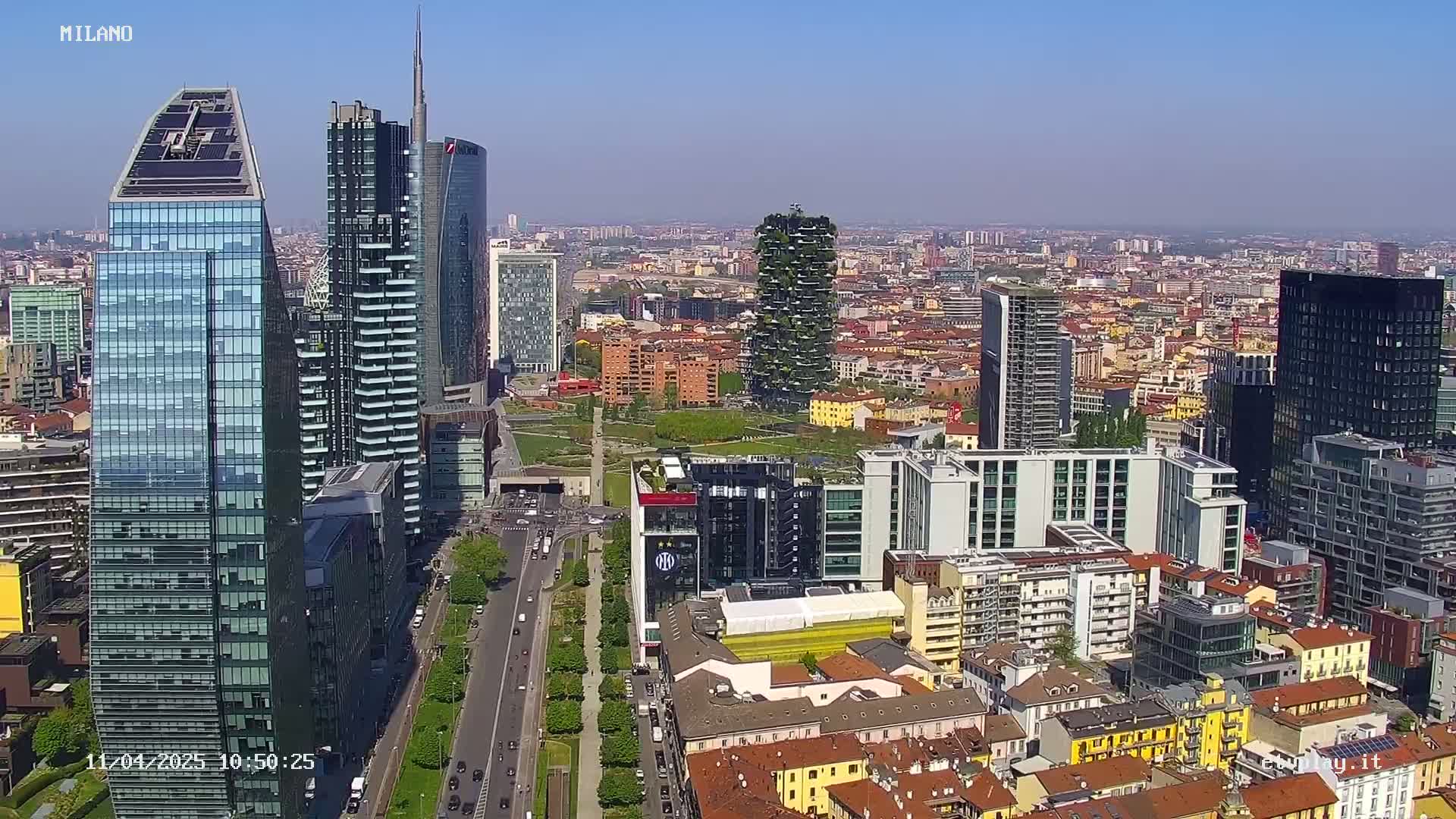 A high-angle, long shot of a dense cityscape on a clear, sunny day, featuring numerous modern skyscrapers and surrounding lower buildings.