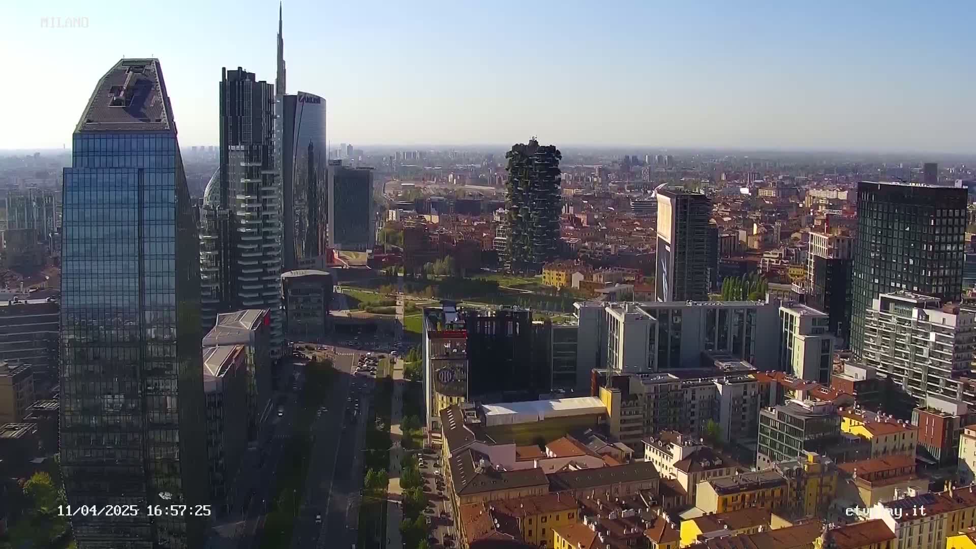 An aerial view of a city on a clear, sunny day shows a mix of modern skyscrapers and traditional low-rise buildings.