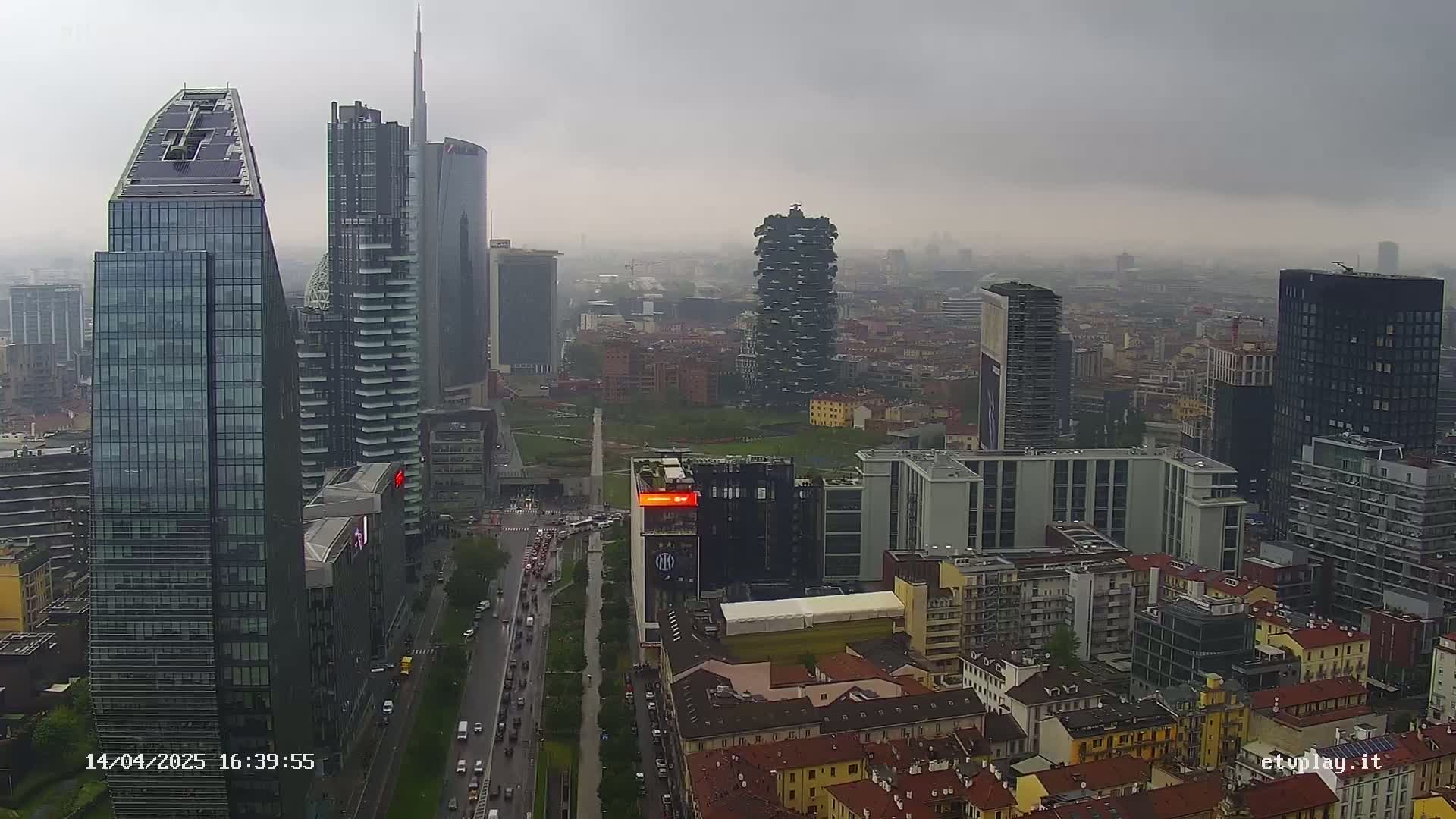 A high-angle view of a city skyline on an overcast day, showing numerous skyscrapers and buildings, with traffic visible on a street below.