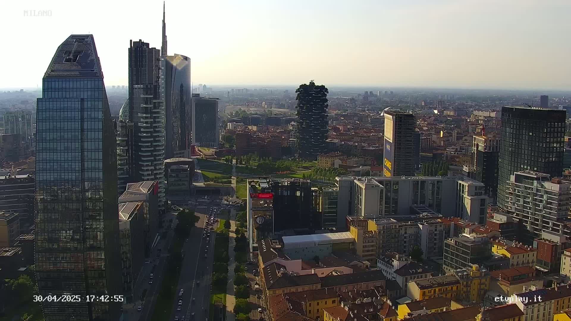 A hazy, sunny day reveals a cityscape of numerous modern and traditional buildings, with several skyscrapers standing tall among a mix of shorter structures and green spaces.